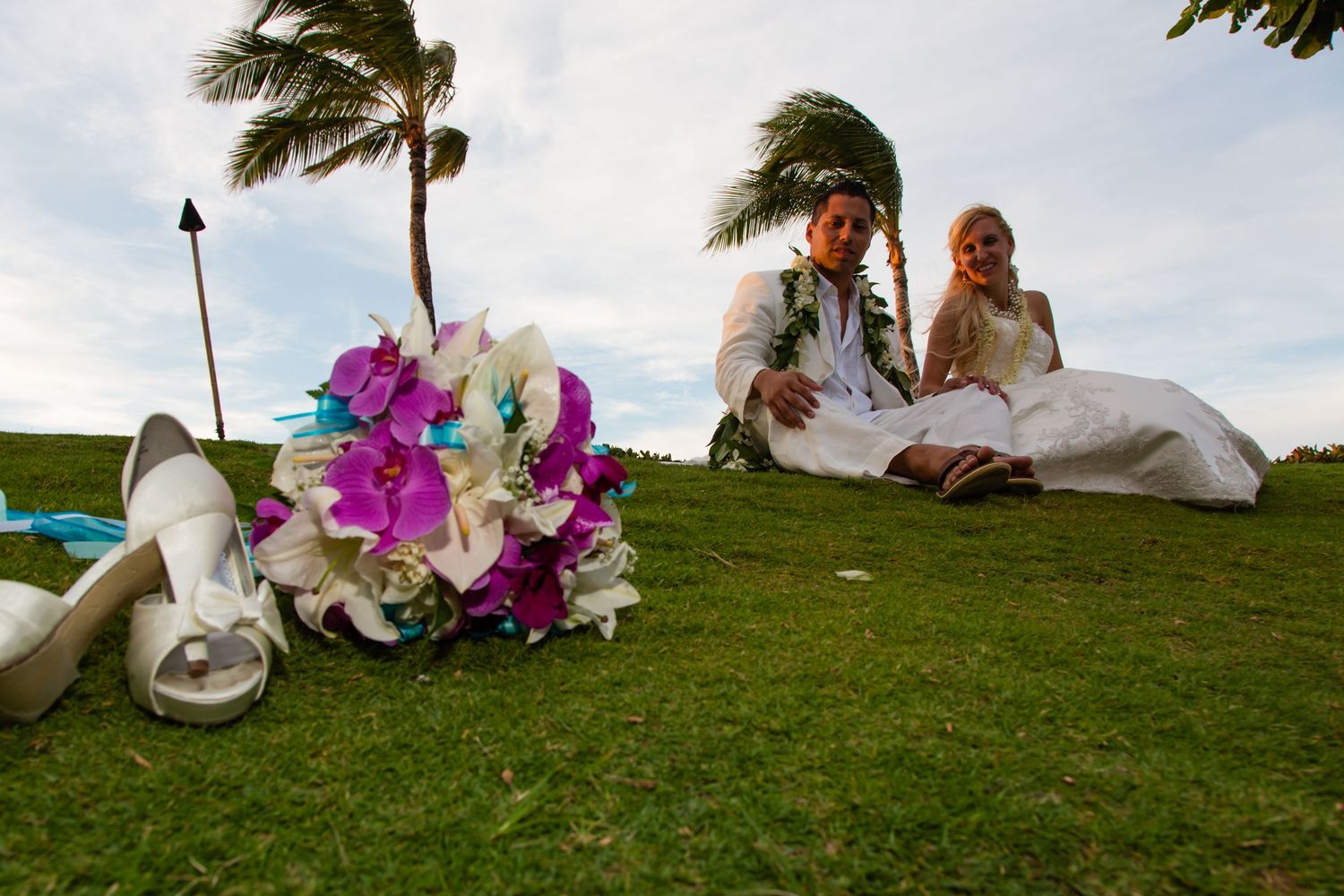 Wedding party members relax on a grassy hillside overlooking the ocean during a beach ceremony in Hawaii.