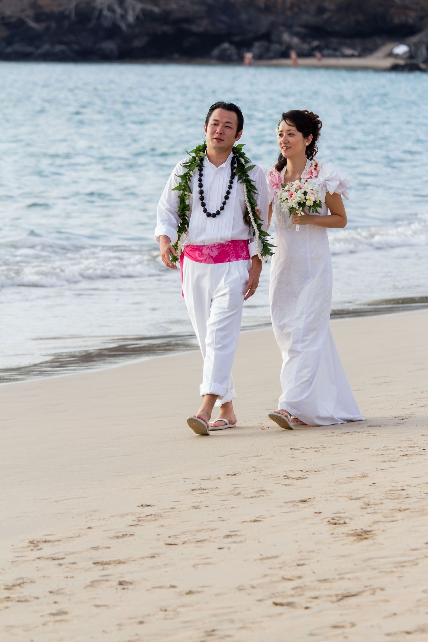 Two people in white attire walk along a sandy beach with ocean waves in the background.