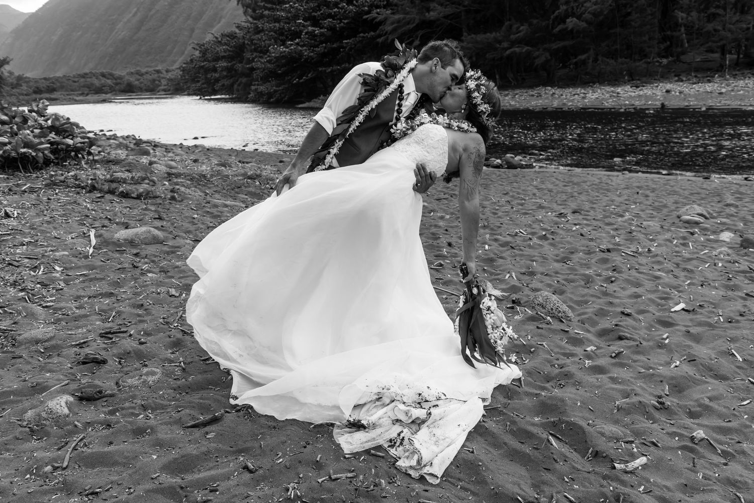 Black-and-white photo of a couple sharing a romantic kiss on a beach in wedding attire, with mountains in the background.