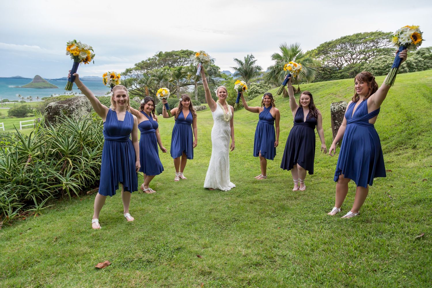 Bridesmaids in royal blue dresses hold yellow bouquets on scenic hilltop.