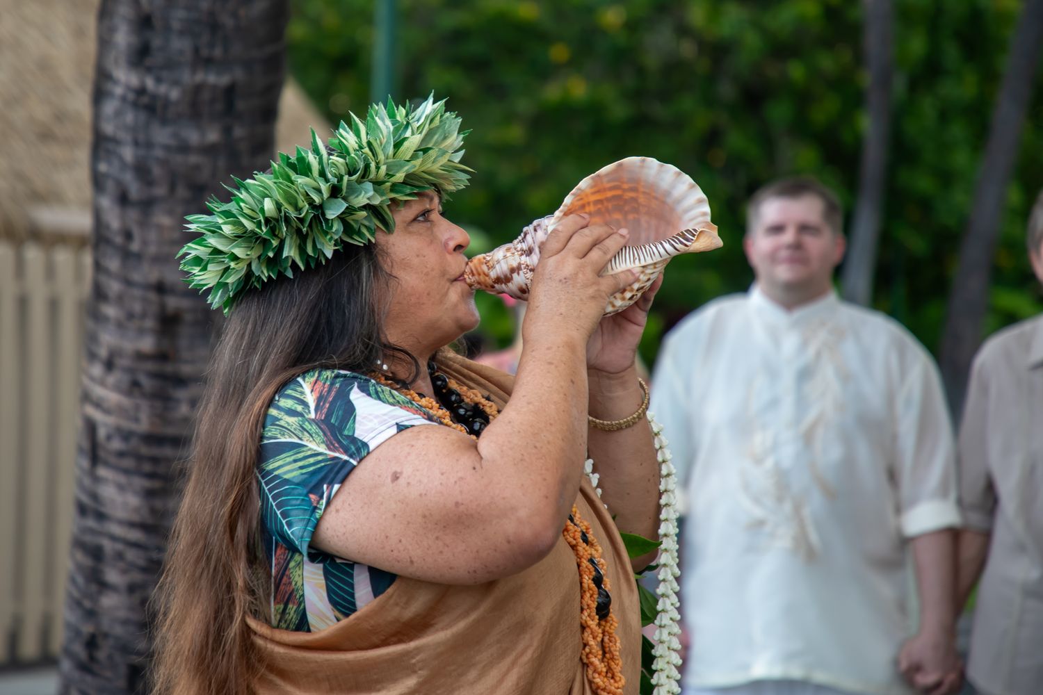 A Hawaiian woman in a leaf crown blows a conch shell trumpet during a traditional cultural ceremony outdoors.