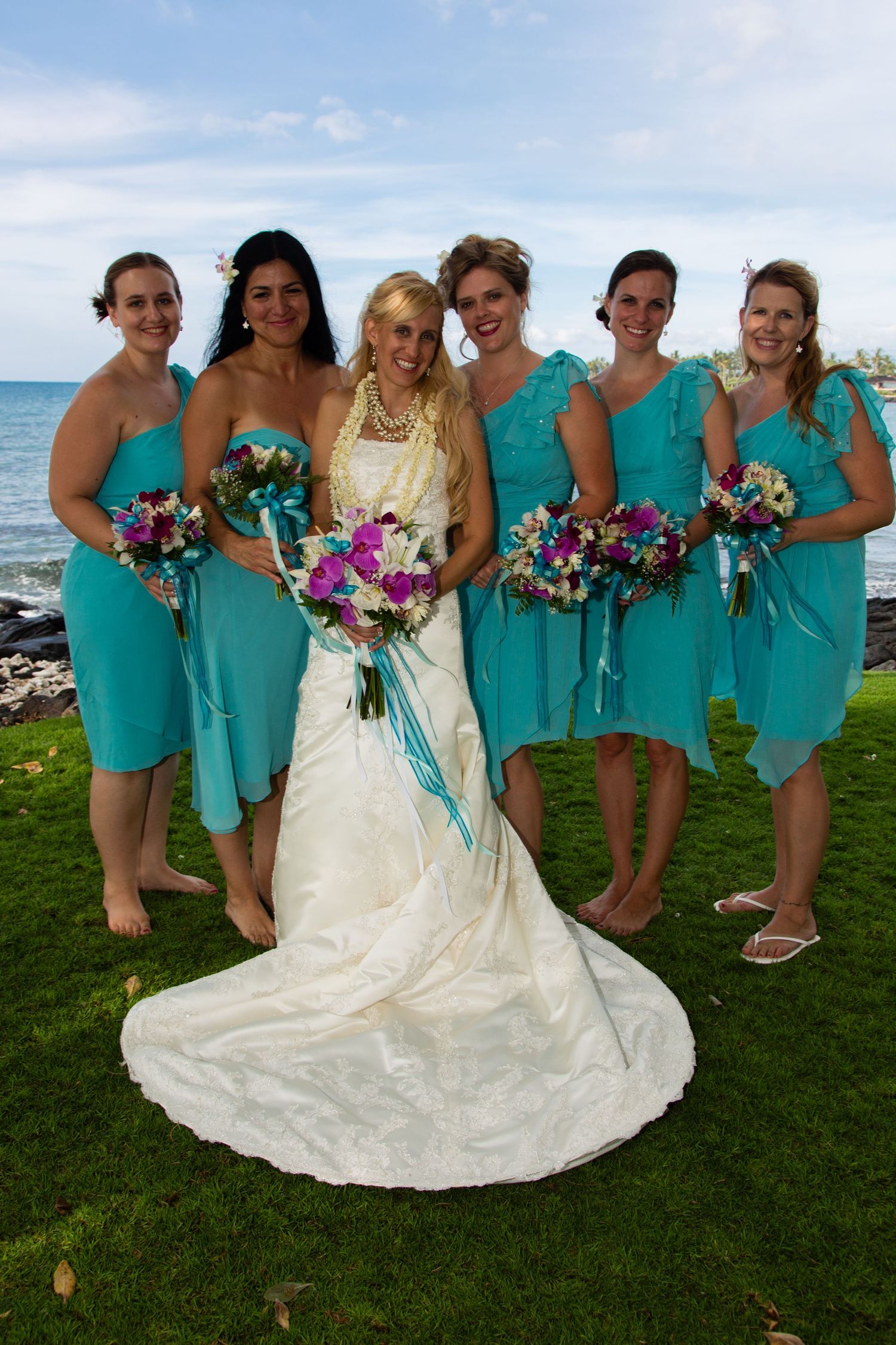 Bridesmaids in turquoise dresses pose with bride in white gown on grassy oceanfront lawn.