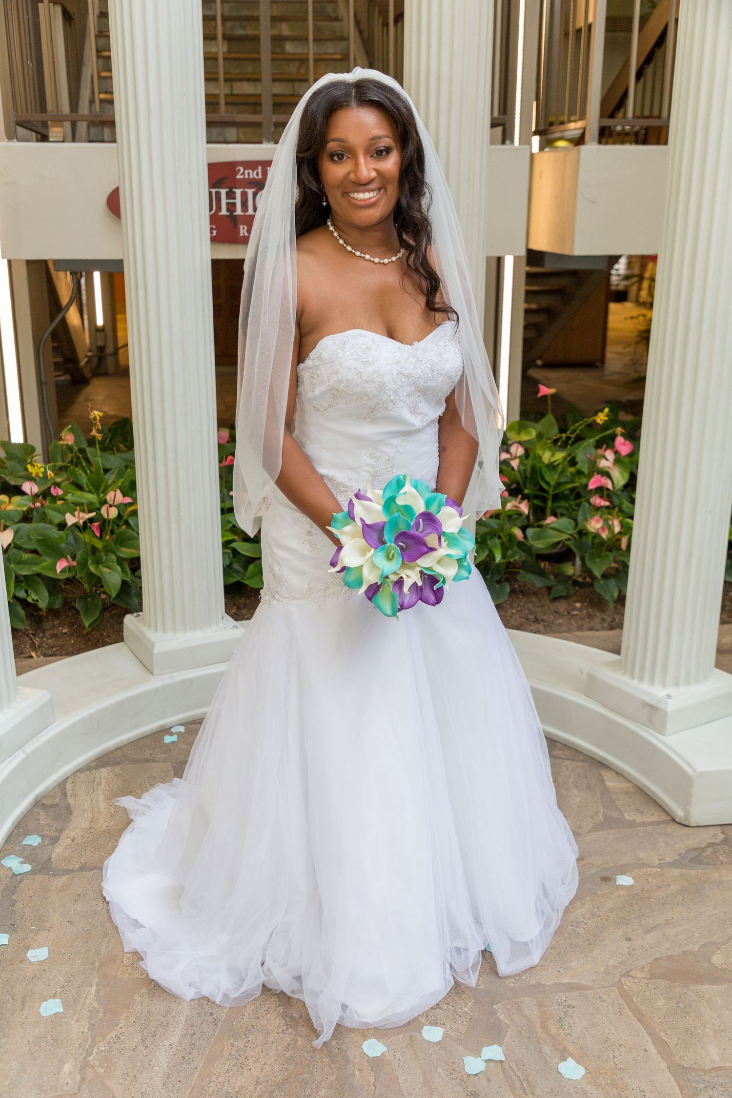 Smiling bride in white strapless gown and veil holds teal and purple bouquet between white columns.