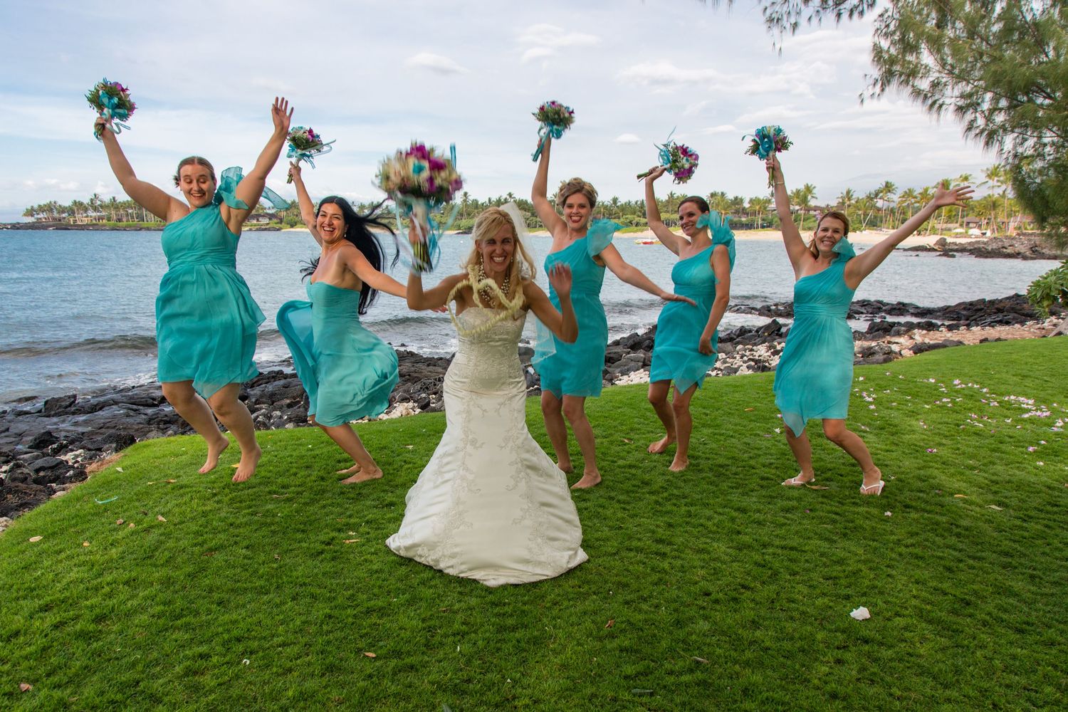 Bridesmaids in turquoise dresses jump joyfully on oceanfront lawn with bride in white gown.