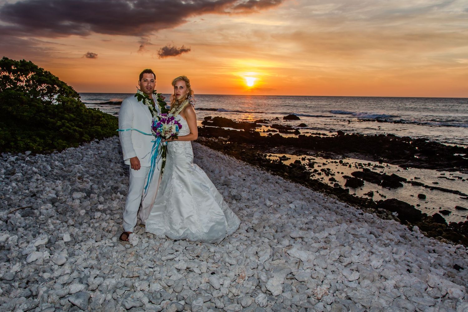 A couple poses on a rocky Hawaiian beach at sunset with orange and pink sky reflecting off the ocean waves.