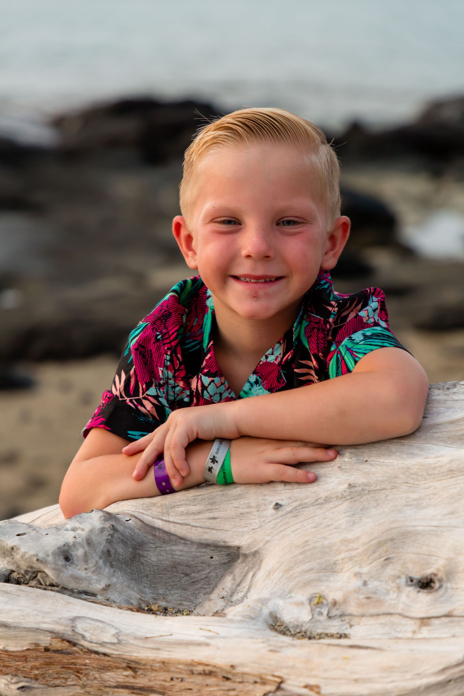 Child in tropical print shirt posing on driftwood at beach during summer photoshoot.
