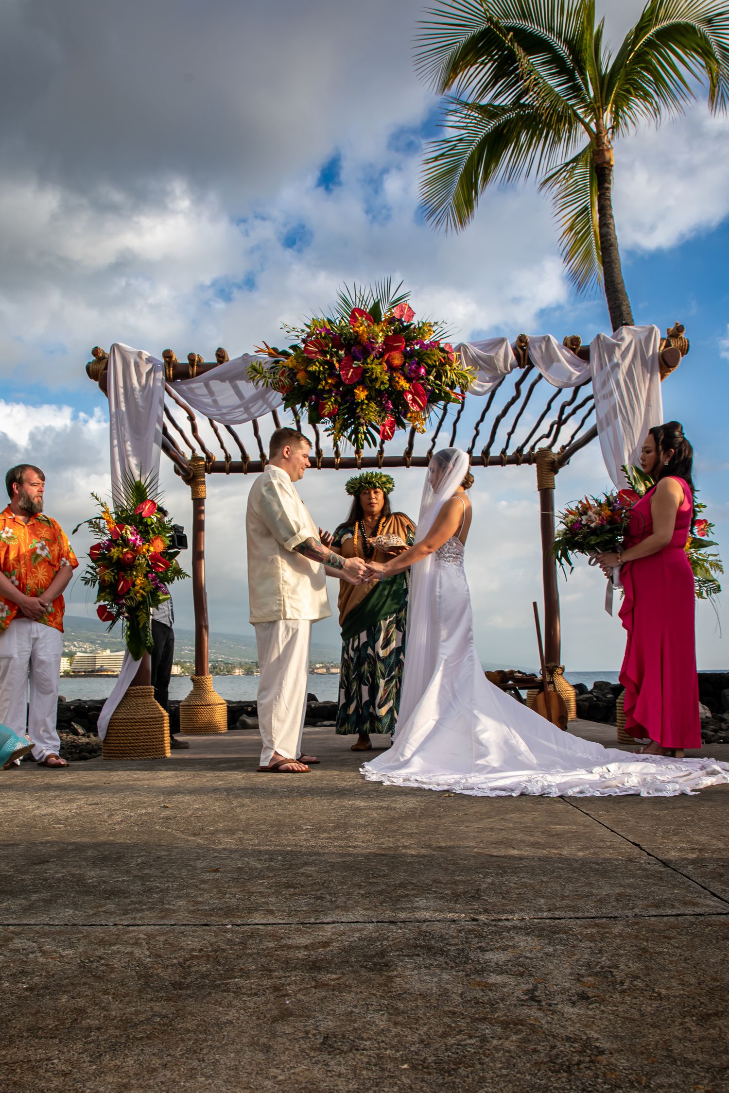 Couple exchanges vows at a Hawaiian beach wedding under a floral arch with a palm tree backdrop.