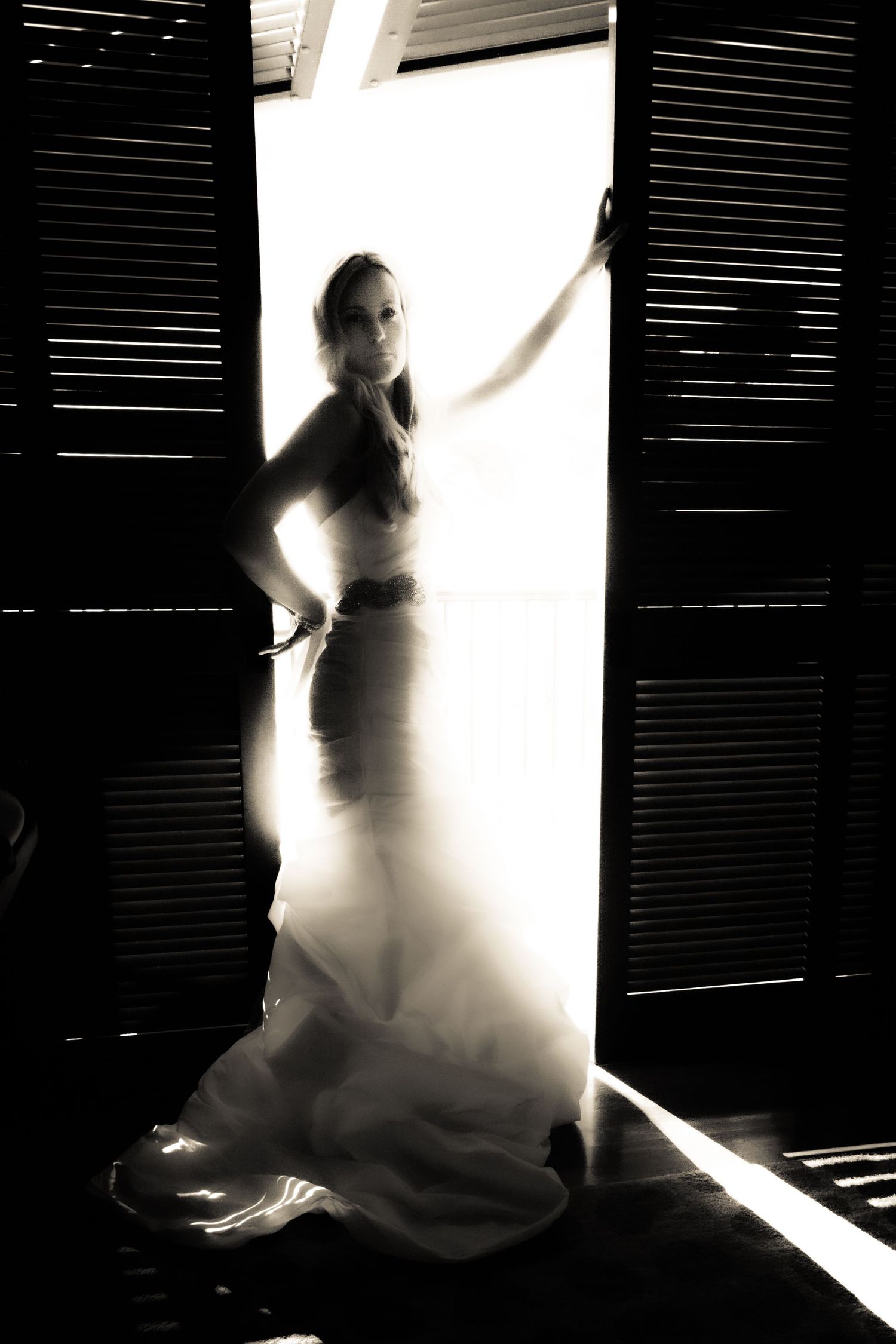 Bride in flowing white wedding gown silhouetted in bright doorway with dark shutters in dramatic black-and-white photo.