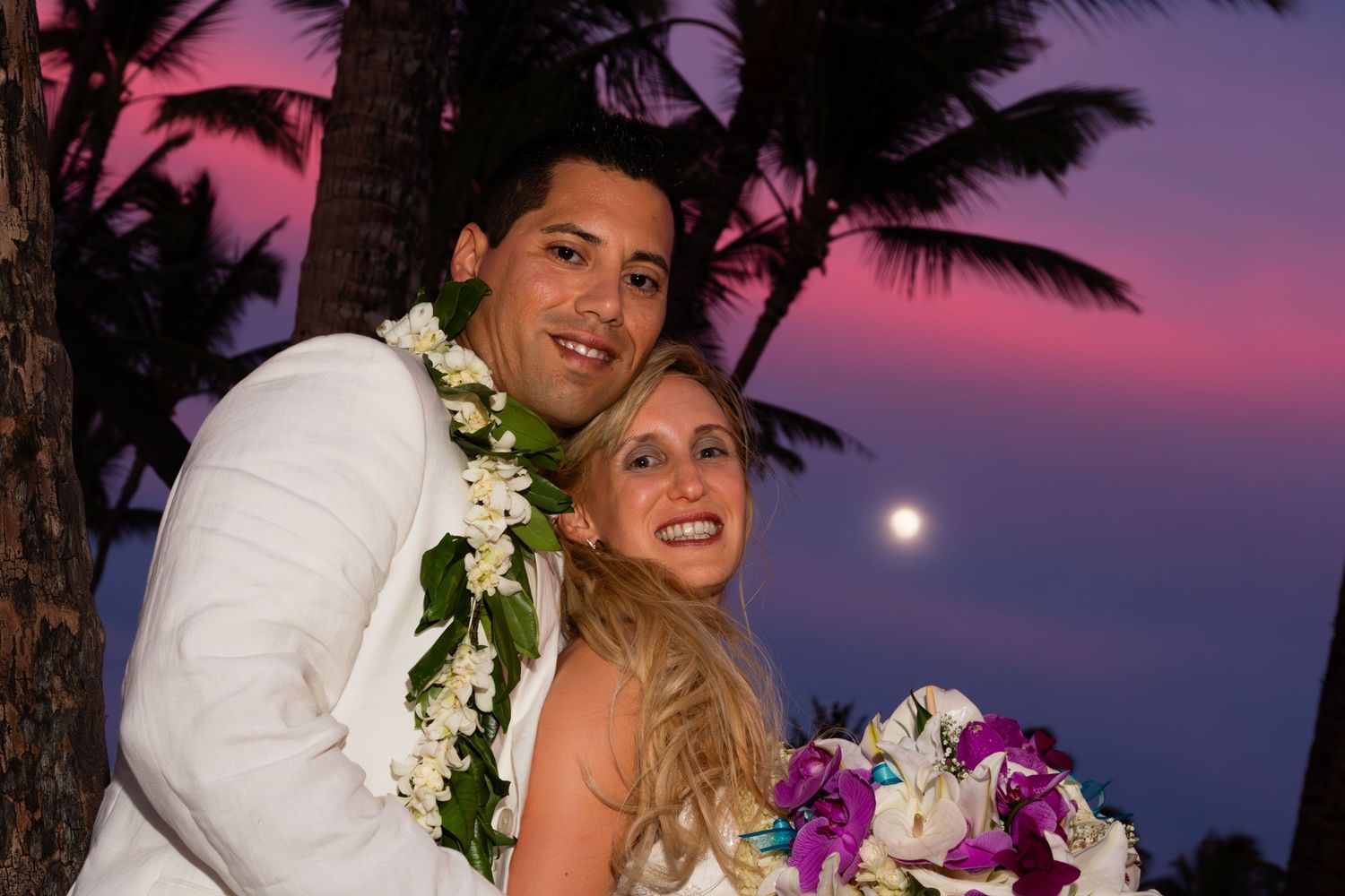 Happy couple shares romantic sunset moment under palm trees at tropical beach wedding with flowers and warm lighting.