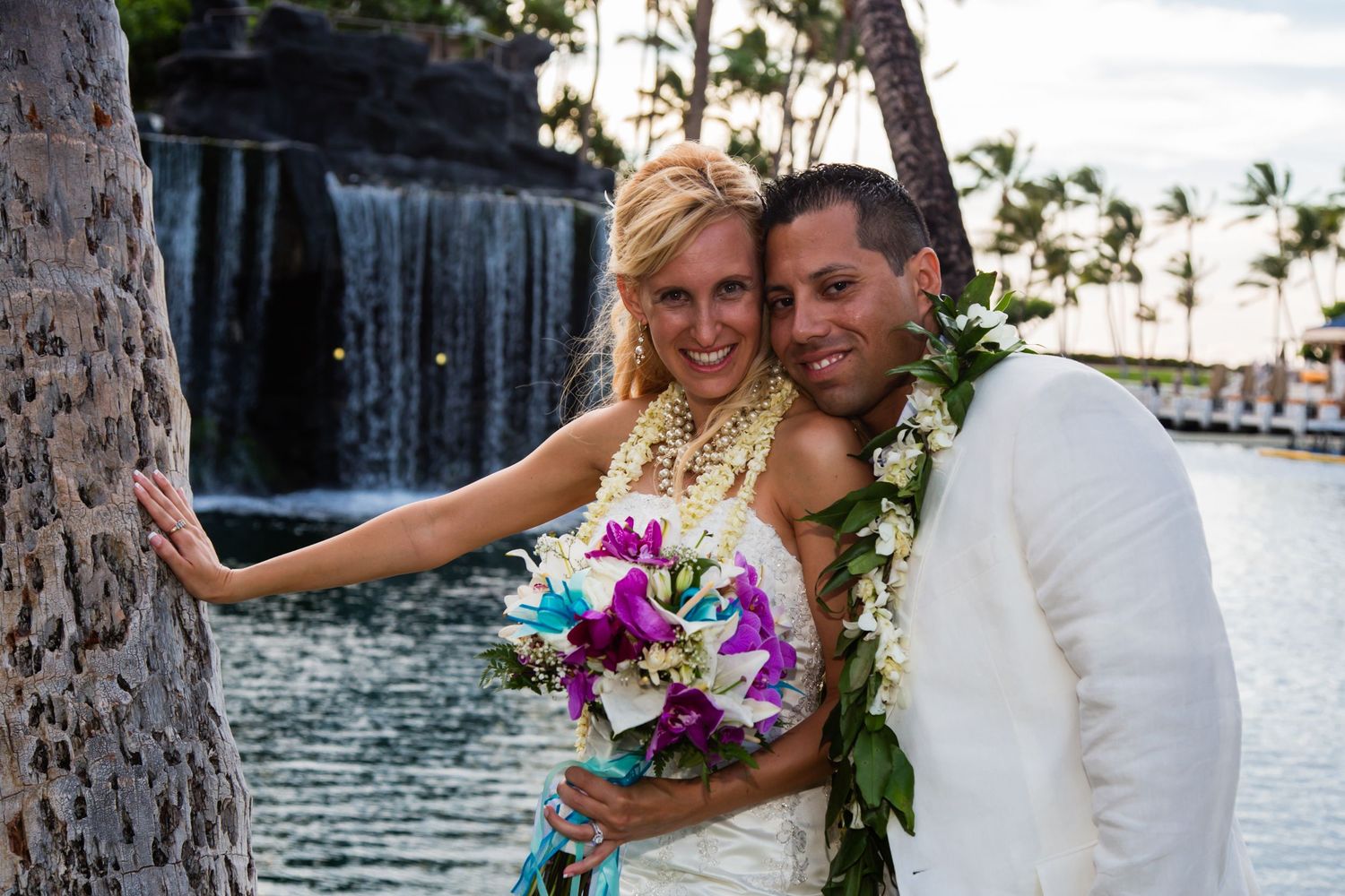 A couple poses for romantic wedding photos in front of a waterfall and tropical palm trees at sunset.