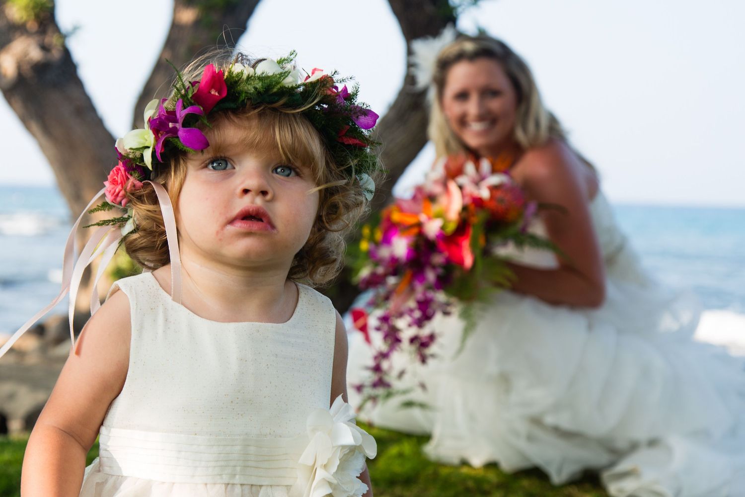 A flower crowned child in white dress stands on beach with wedding dress in background at Kikaua Beach Park in Kailua Kona, Hawaii