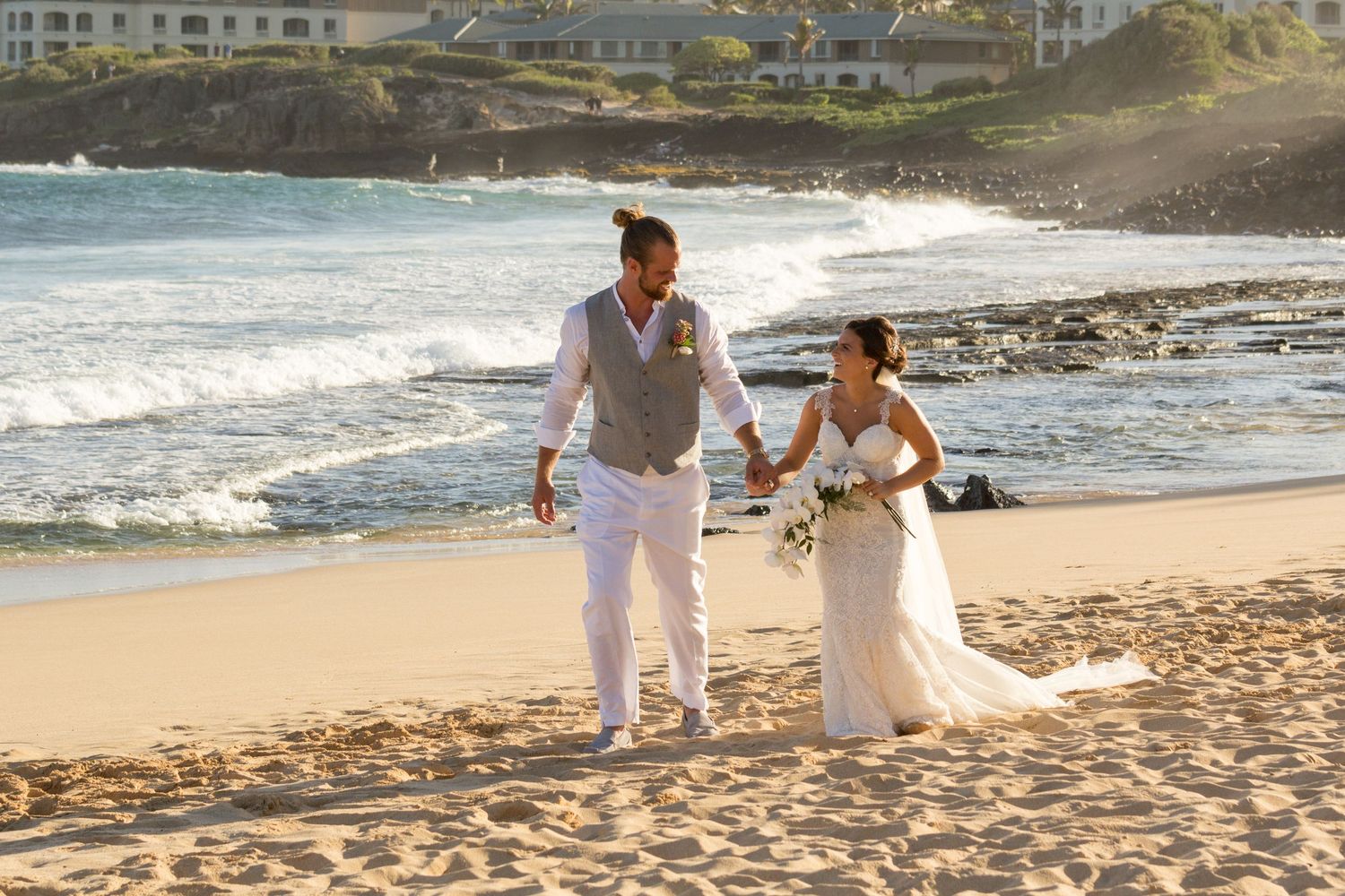 Newlywed couple walking hand in hand on a sunny beach, bride in white gown, groom in vest and white pants.