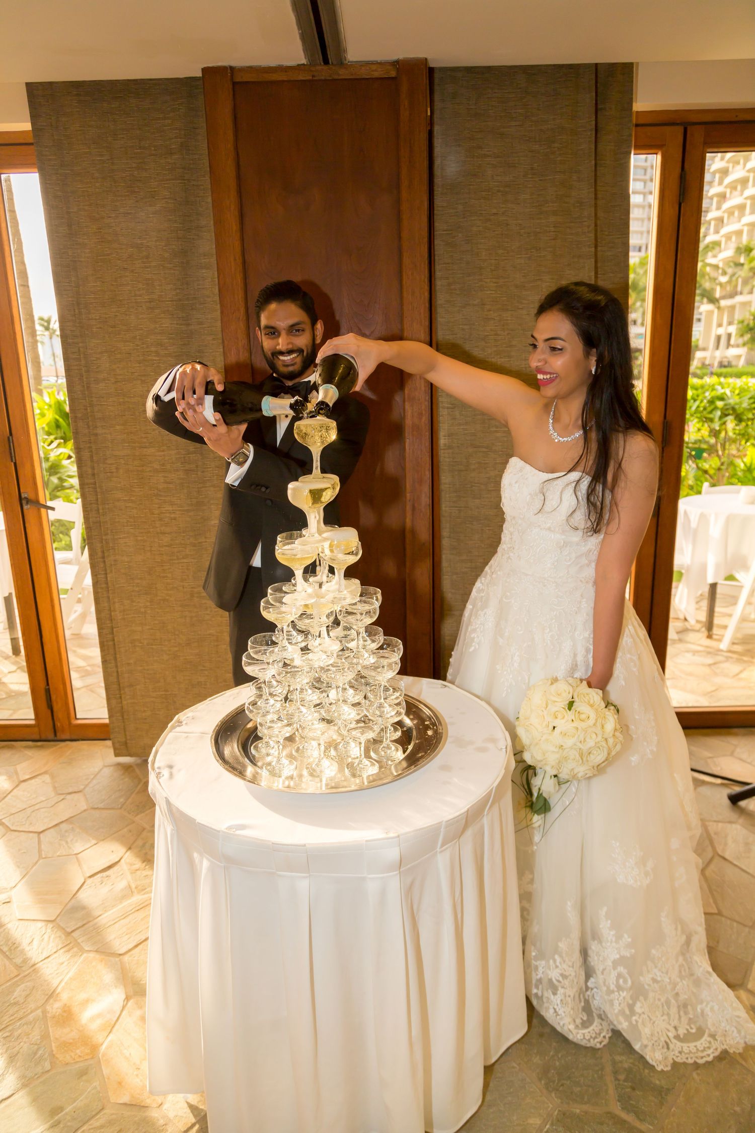 Bride and groom pouring champagne tower at wedding reception, bride holding white bouquet in elegant lace gown.