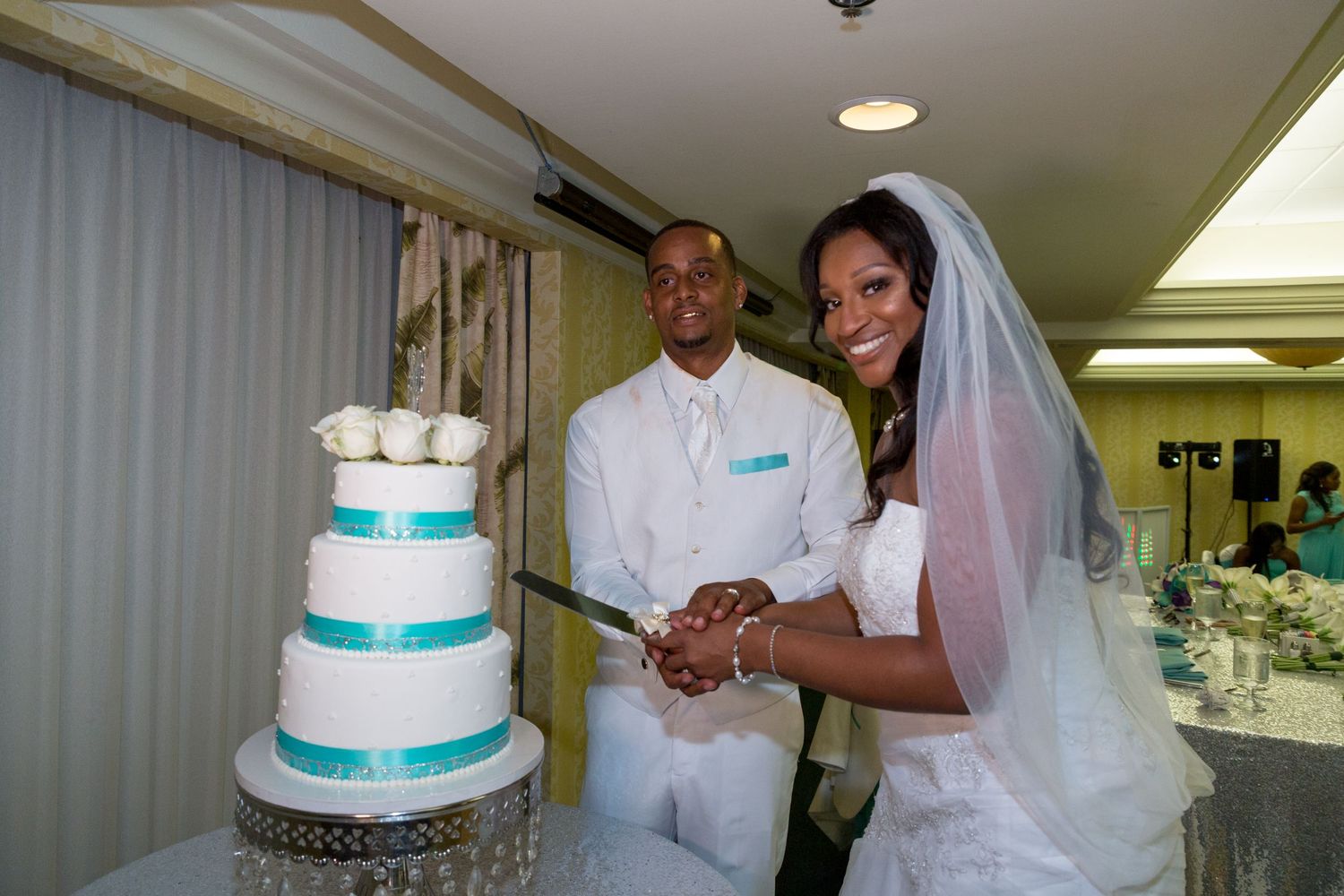 Couple cutting elegant three-tiered white and turquoise wedding cake at indoor reception.