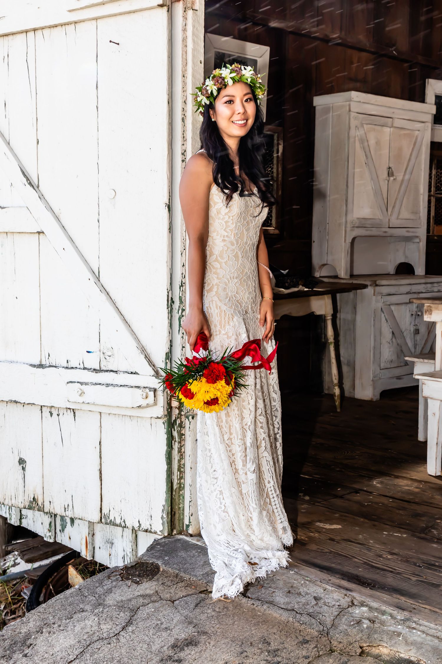 A bride in a lace dress and flower crown poses in a rustic doorway holding a colorful bouquet photographed in Waimea, Hawaii.