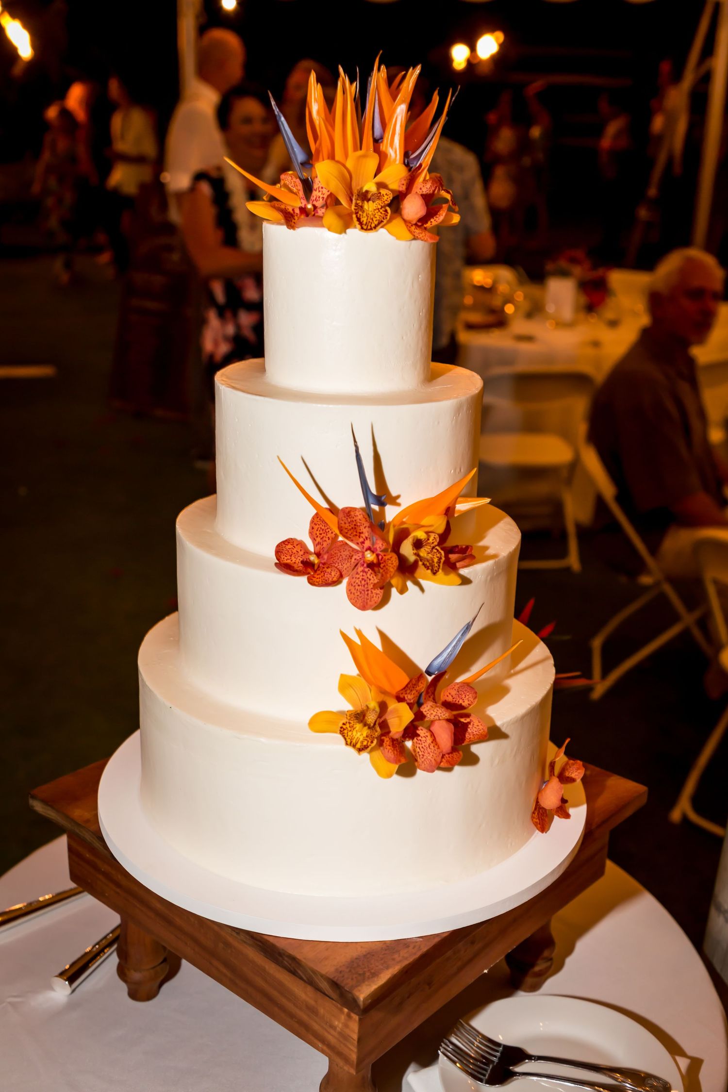 Elegant four-tier white wedding cake adorned with vibrant orange bird of paradise flowers on a wooden stand at a reception.