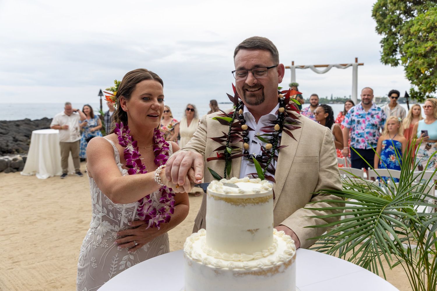 Bride and groom cutting white wedding cake on beach in Hawaii, wearing leis, with guests in background.