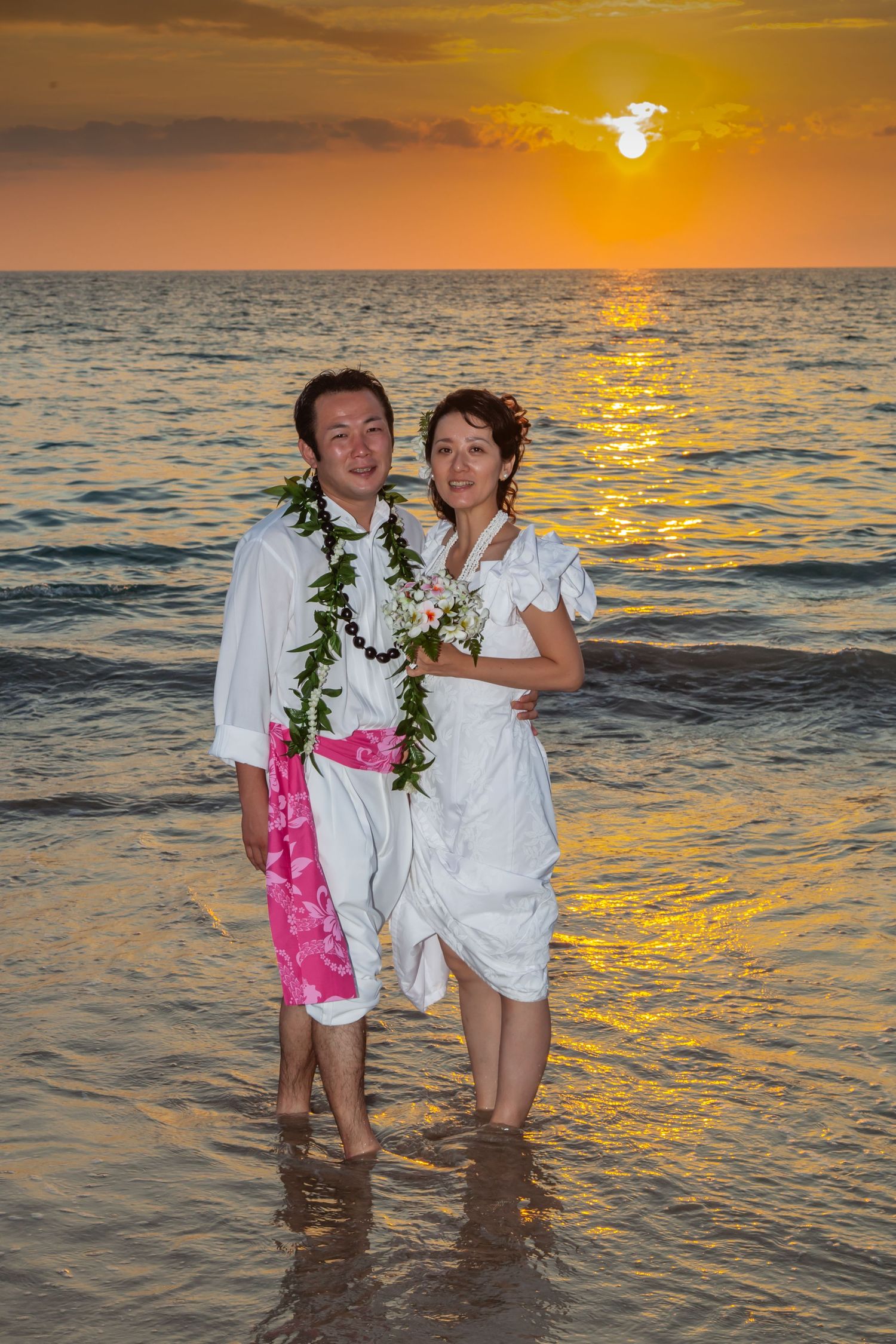 Couple wearing white stands in ocean water during golden sunset creating romantic silhouettes against orange sky.
