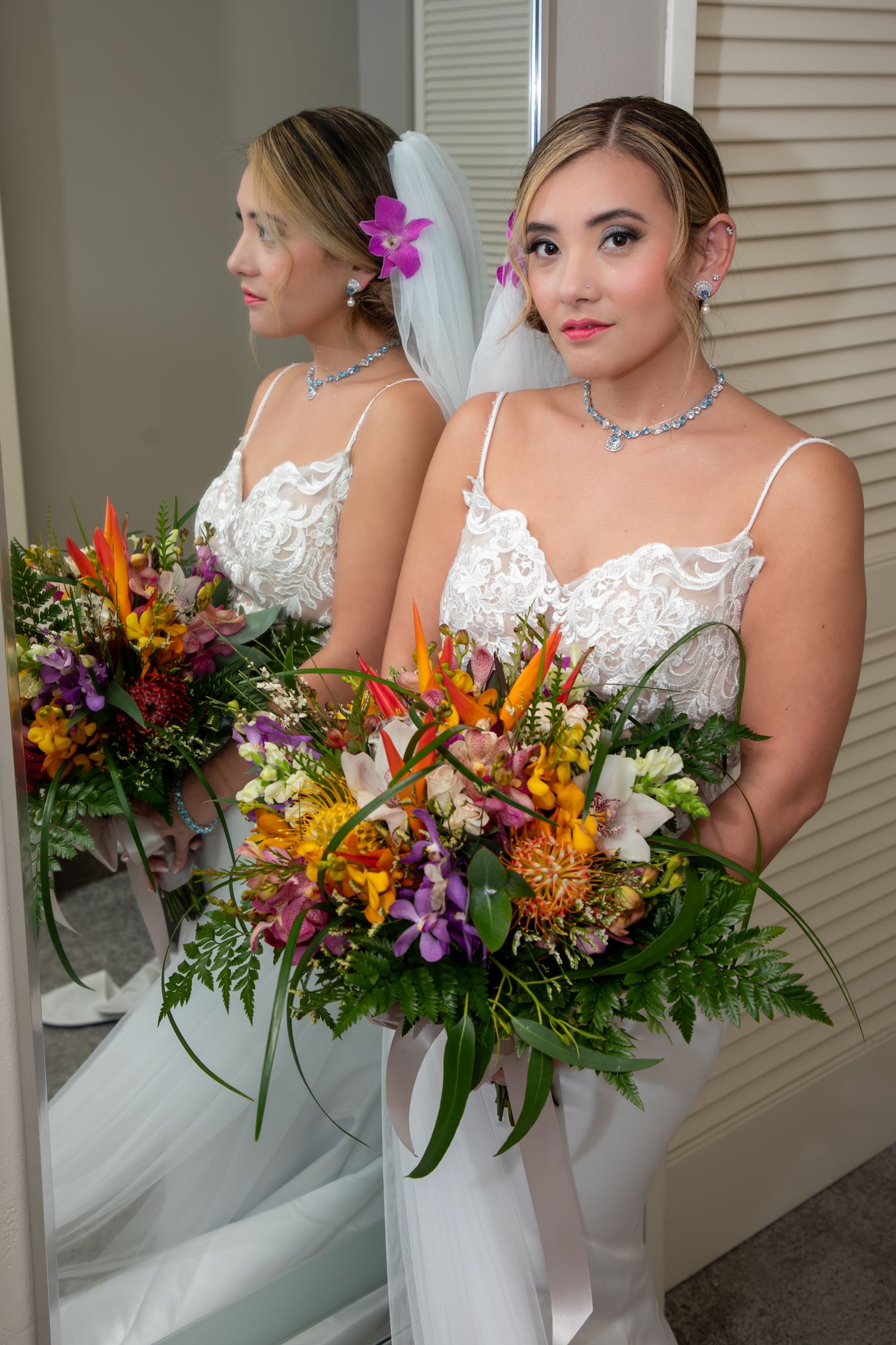 A bride in a white spaghetti strap lace gown poses with a vibrant fall-colored bouquet next to a mirror reflection.