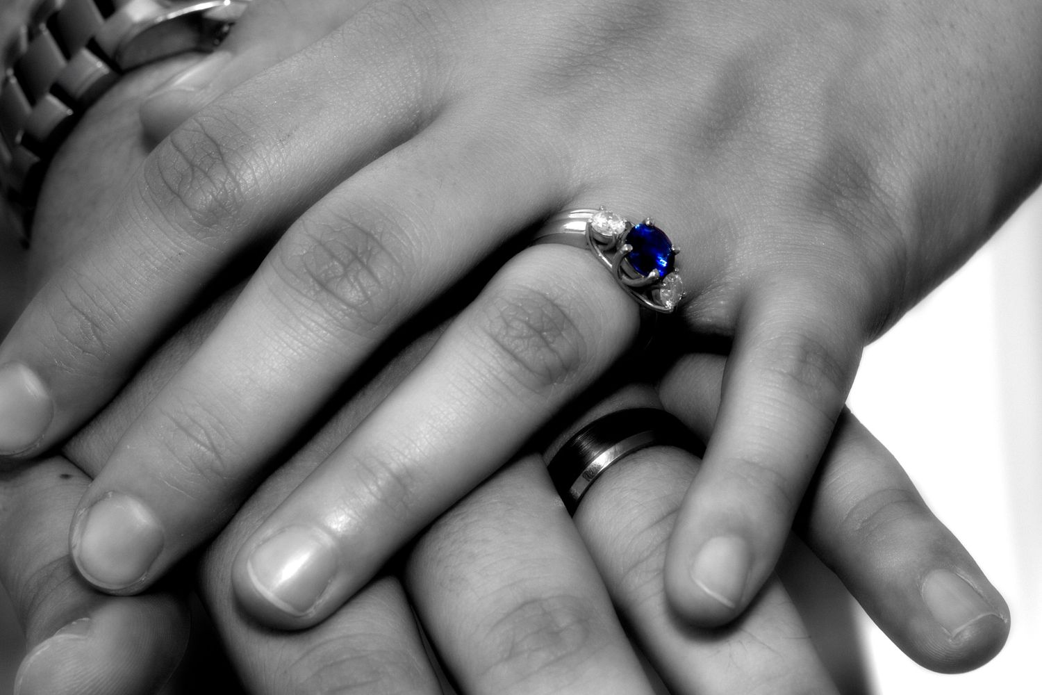 Black and white photo of intertwined hands with a sapphire ring prominently displayed.