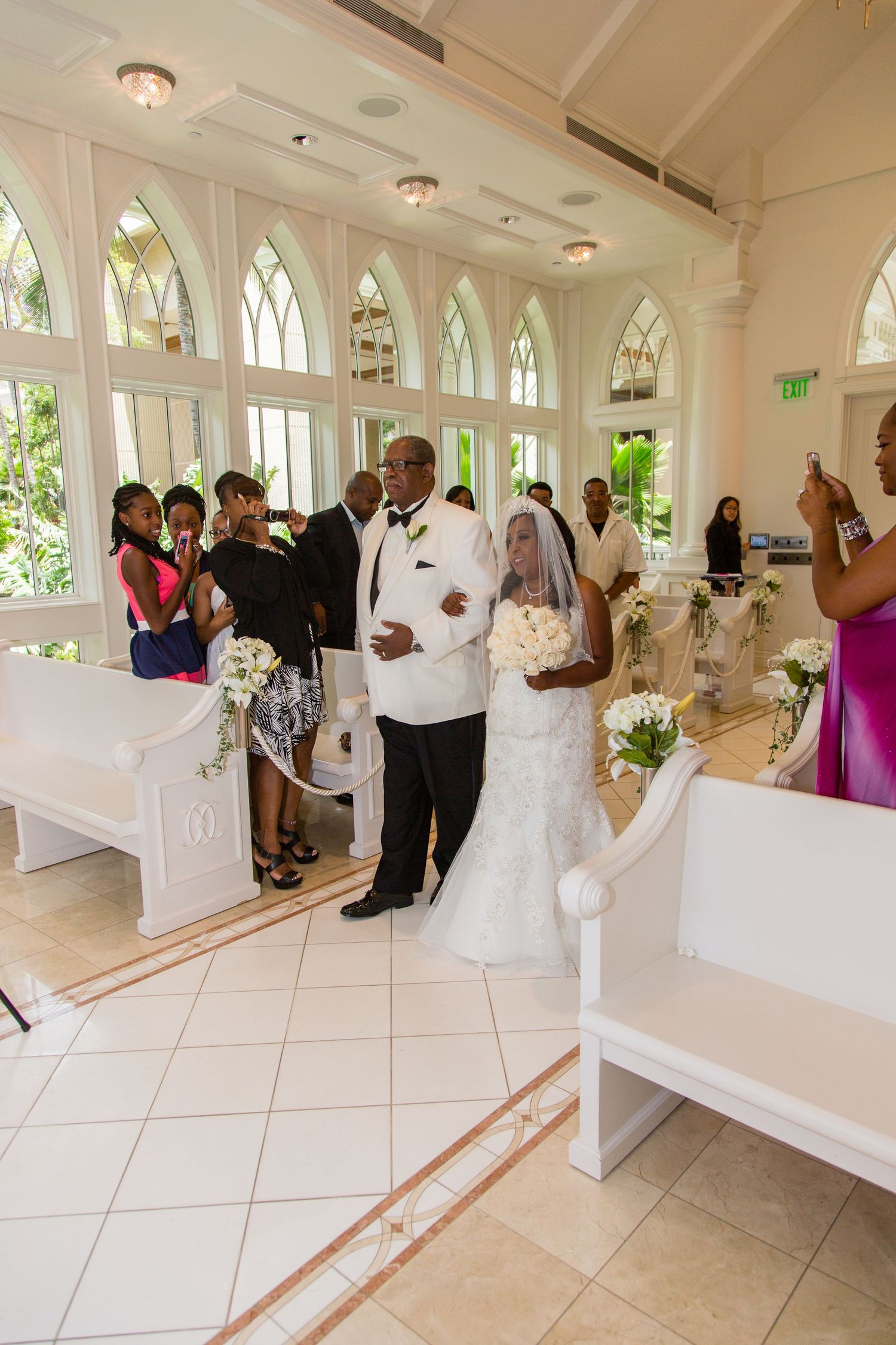 Bride walks down the aisle with her father in a bright white chapel during a wedding ceremony.