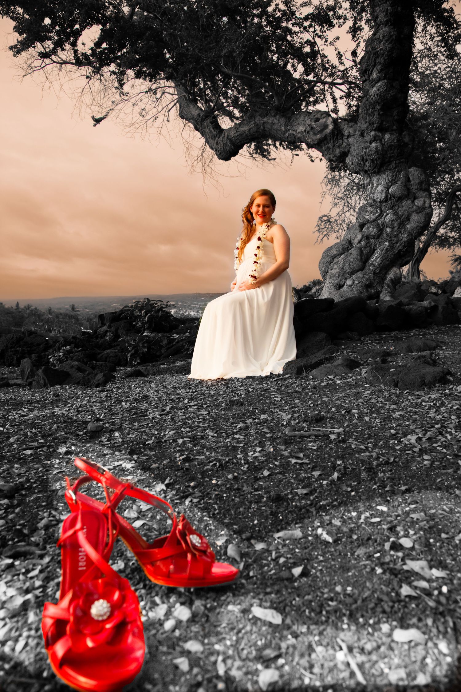 Red wedding shoes in foreground with bride seated under tree in selective color photo.