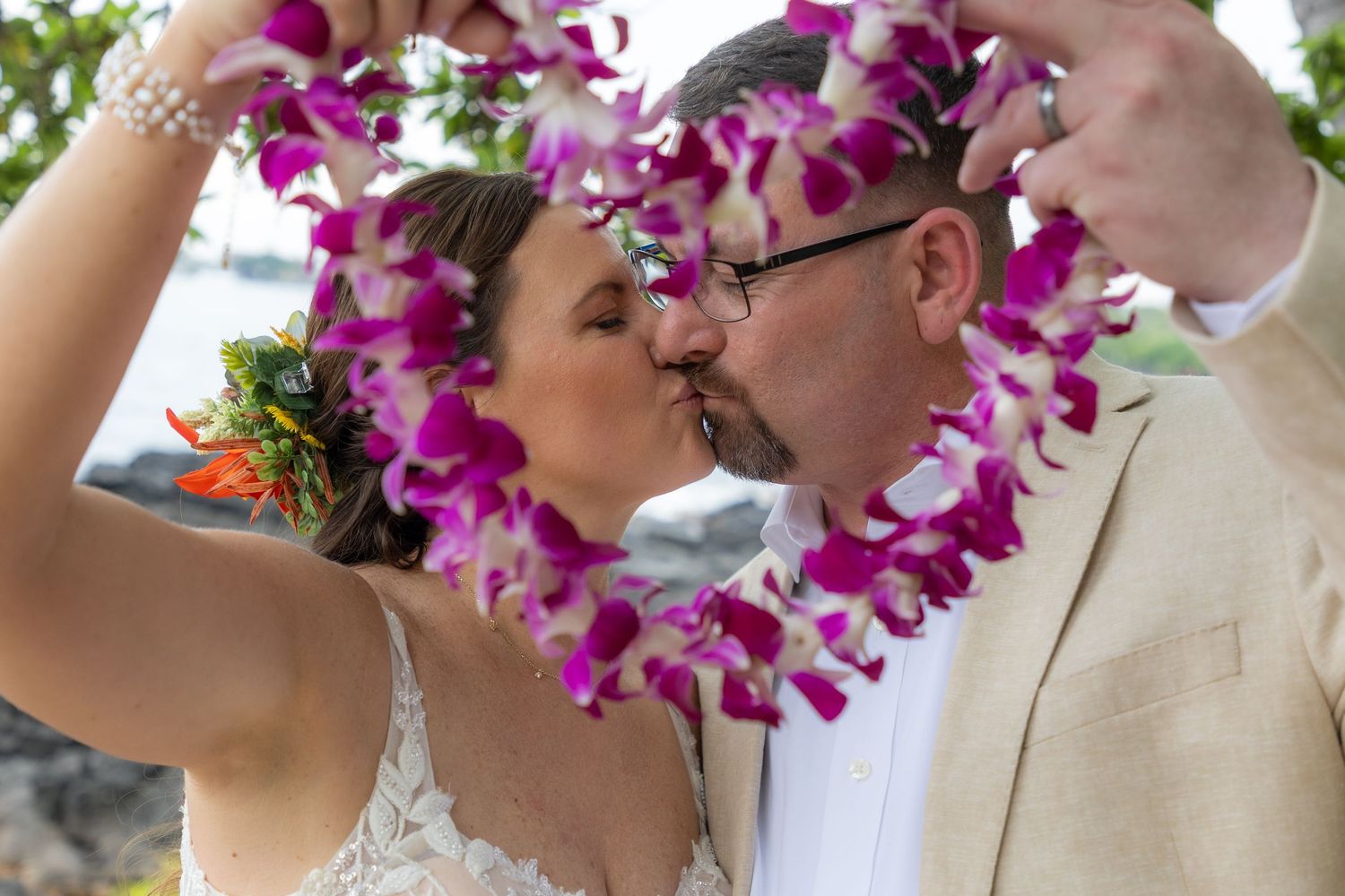 Newlywed couple kissing framed by purple orchid leis during outdoor Hawaiian wedding ceremony.