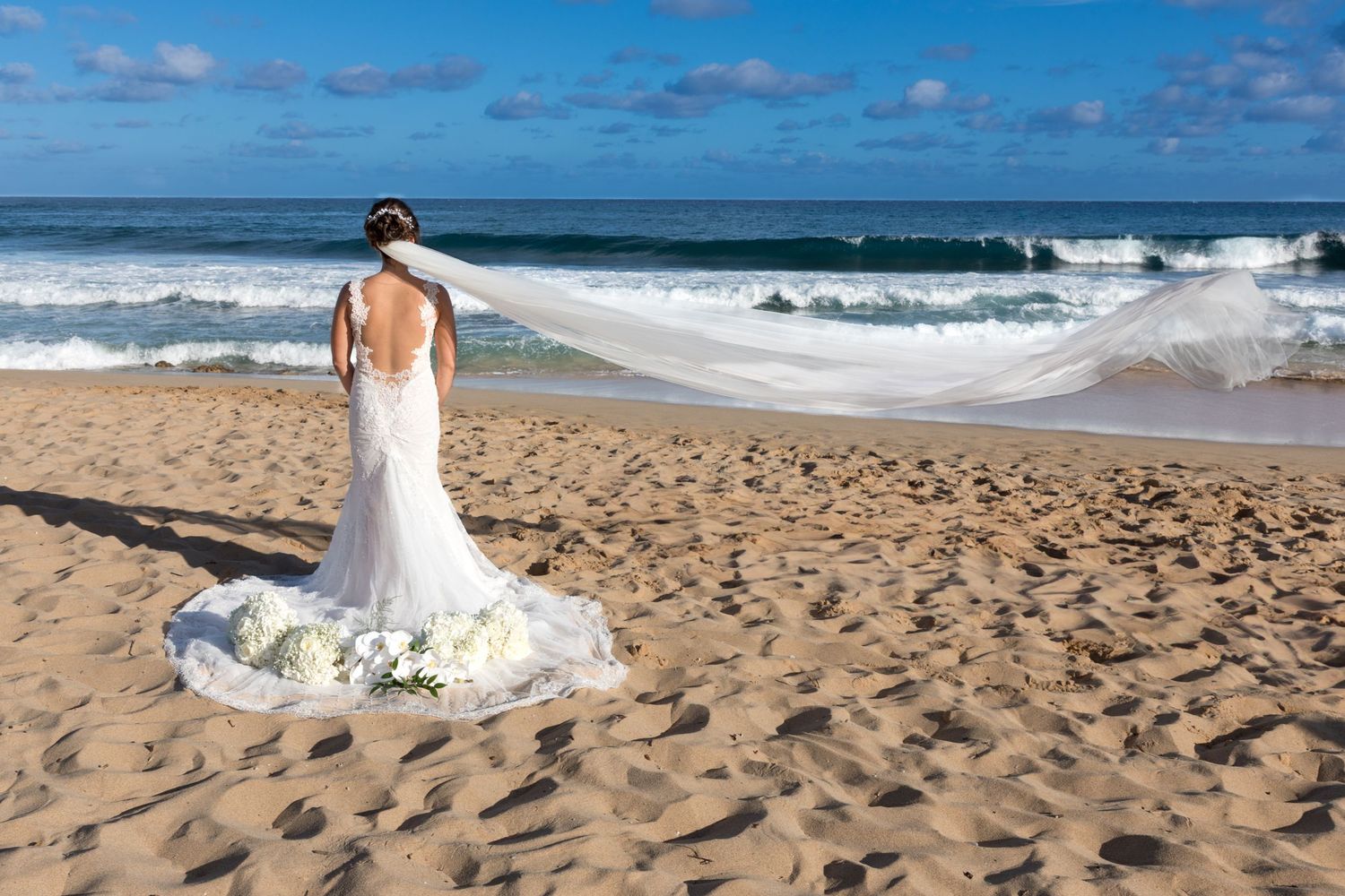 Back view of bride in fitted lace dress standing on sandy beach watching waves in Kauai.