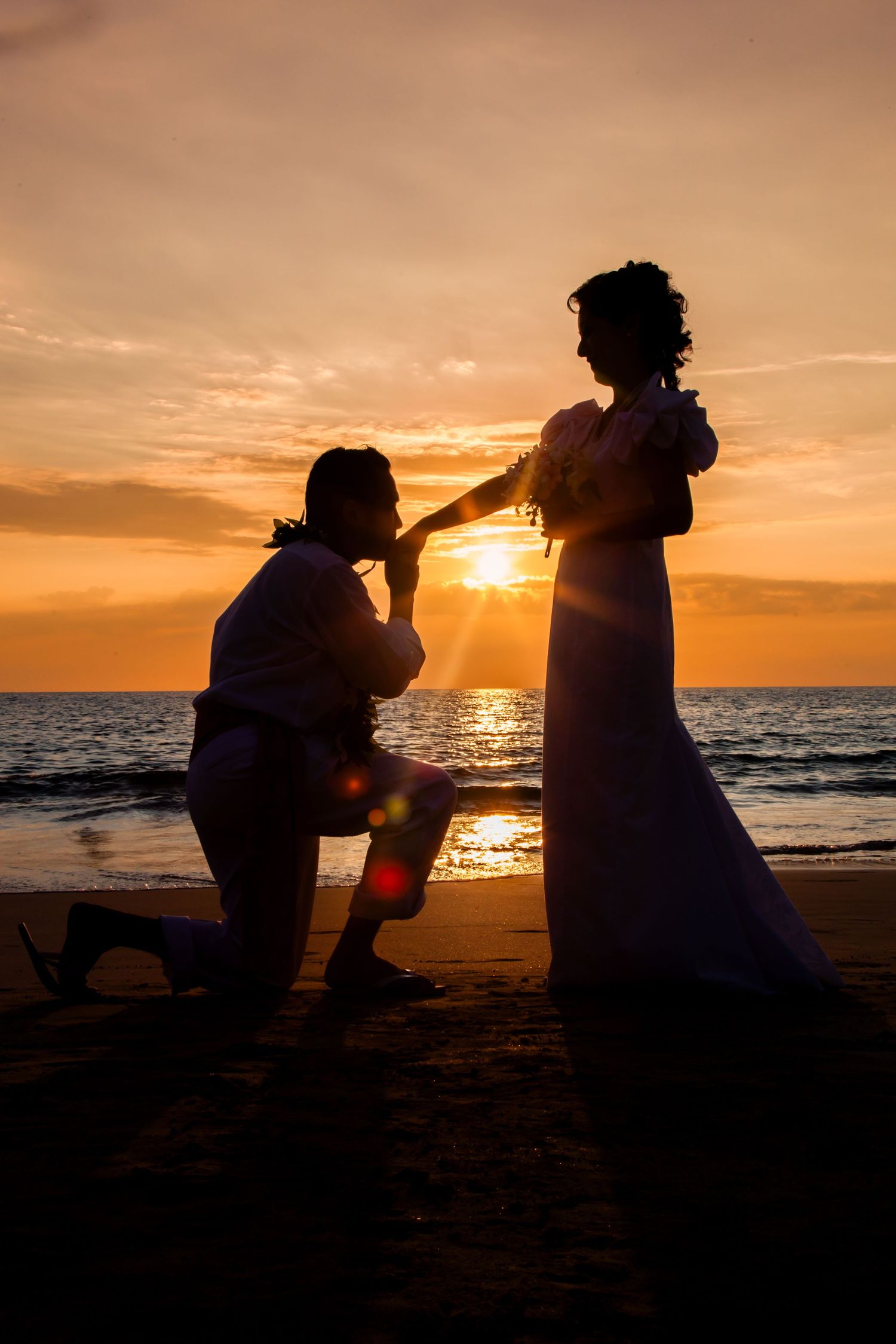 Hawaiian Sunset Photography captures a silhouetted groom kissing his bride's hand at a Kailua-Kona beach sunset. *