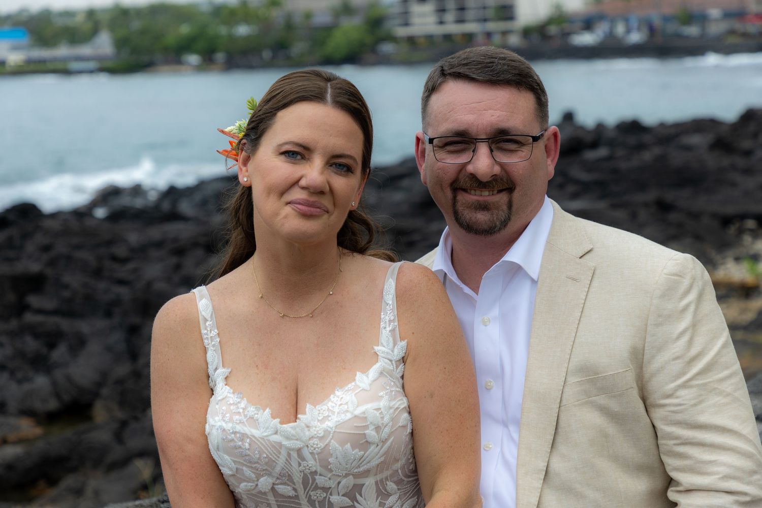 Couple in wedding attire smiling together on rocky Hawaiian coastline with ocean views.