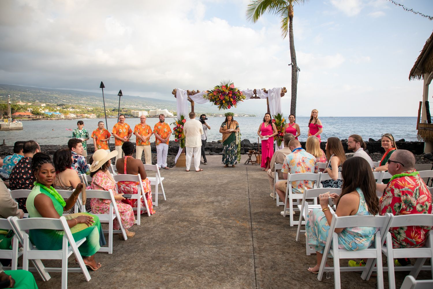 Outdoor Hawaiian wedding ceremony by the ocean with guests seated on white chairs under a colorful floral arch.