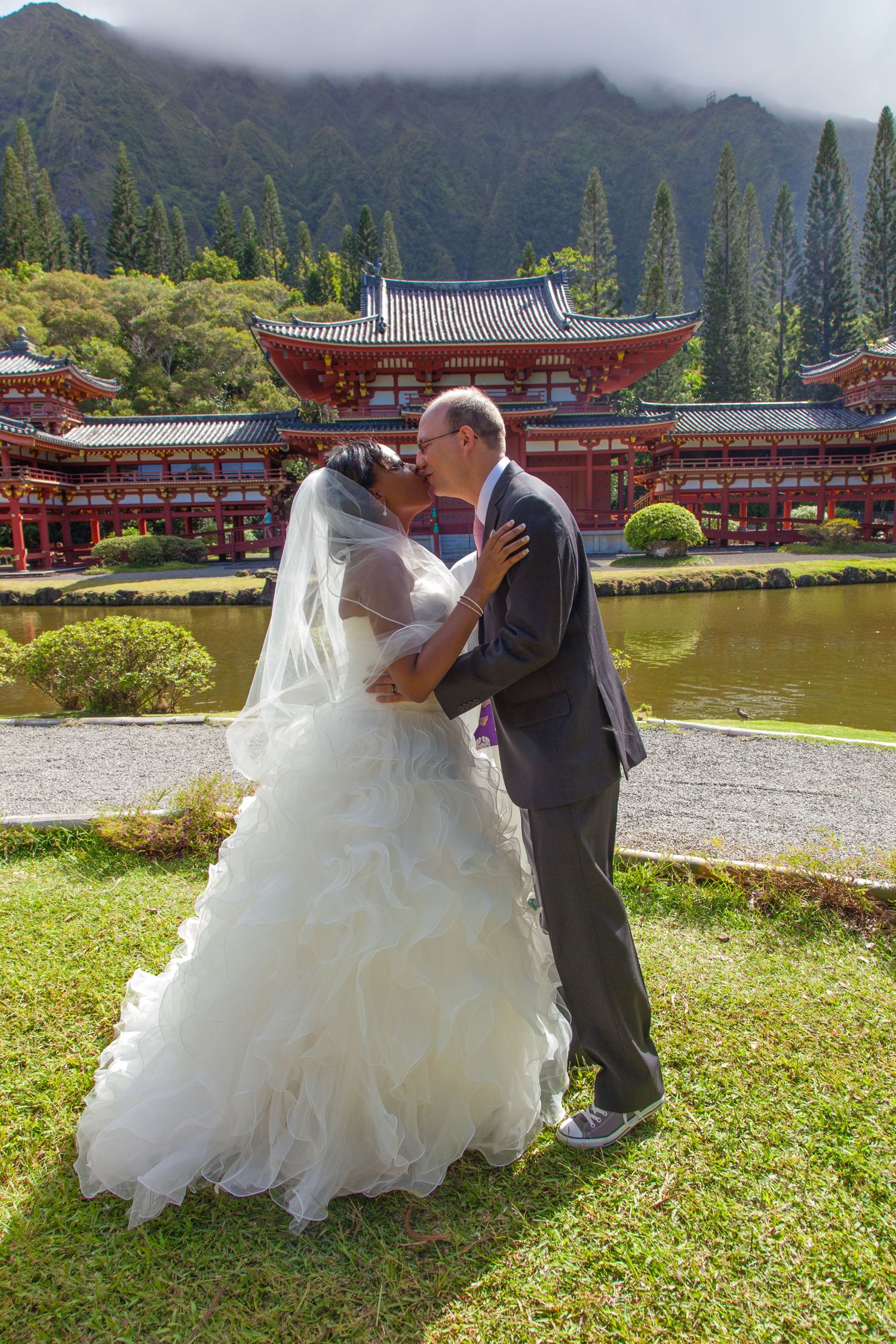 A couple shares romantic moments in traditional wedding attire by a serene lake with Japanese temple buildings in the background.