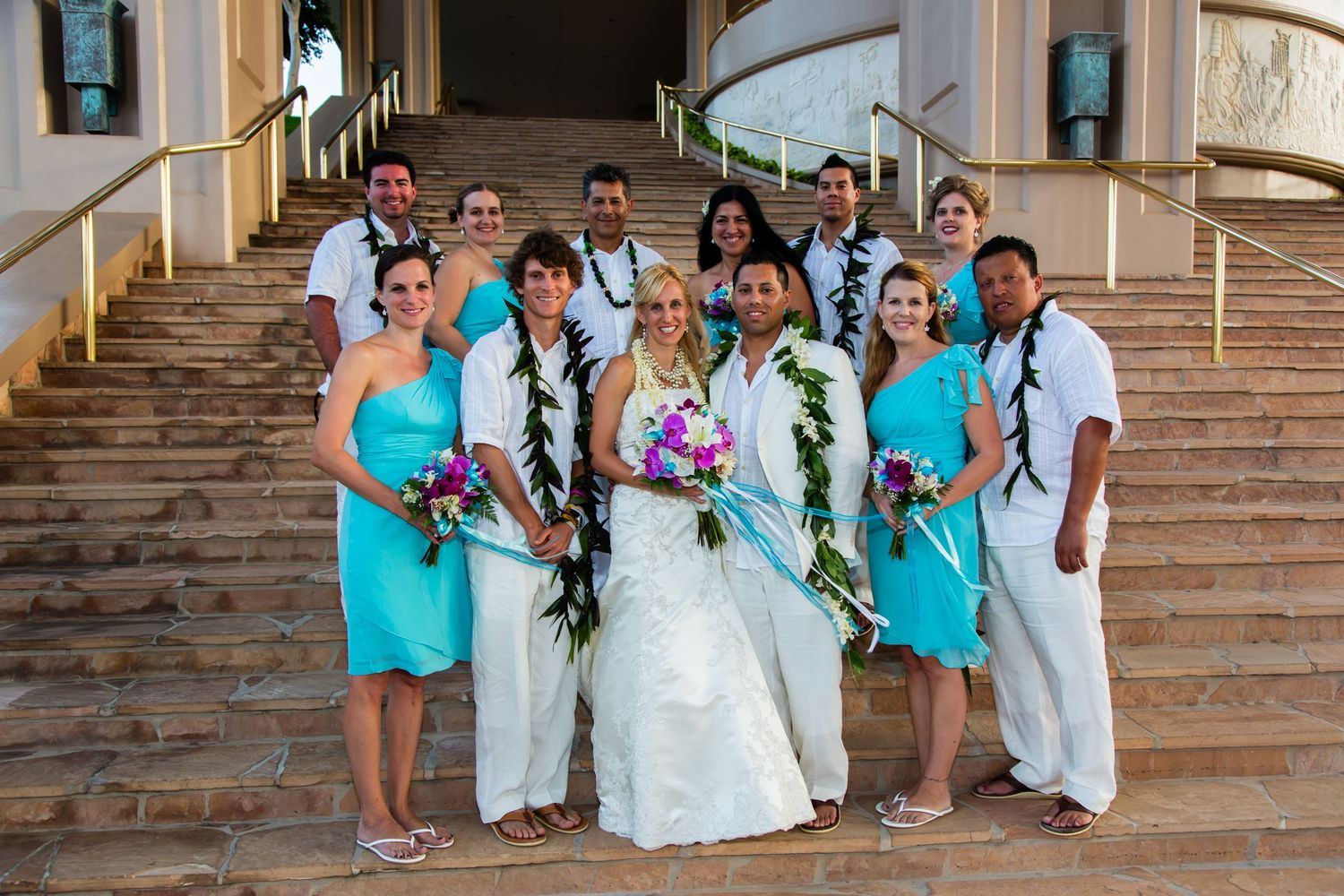 Wedding party members in turquoise and white attire pose together on stone steps during an outdoor celebration.