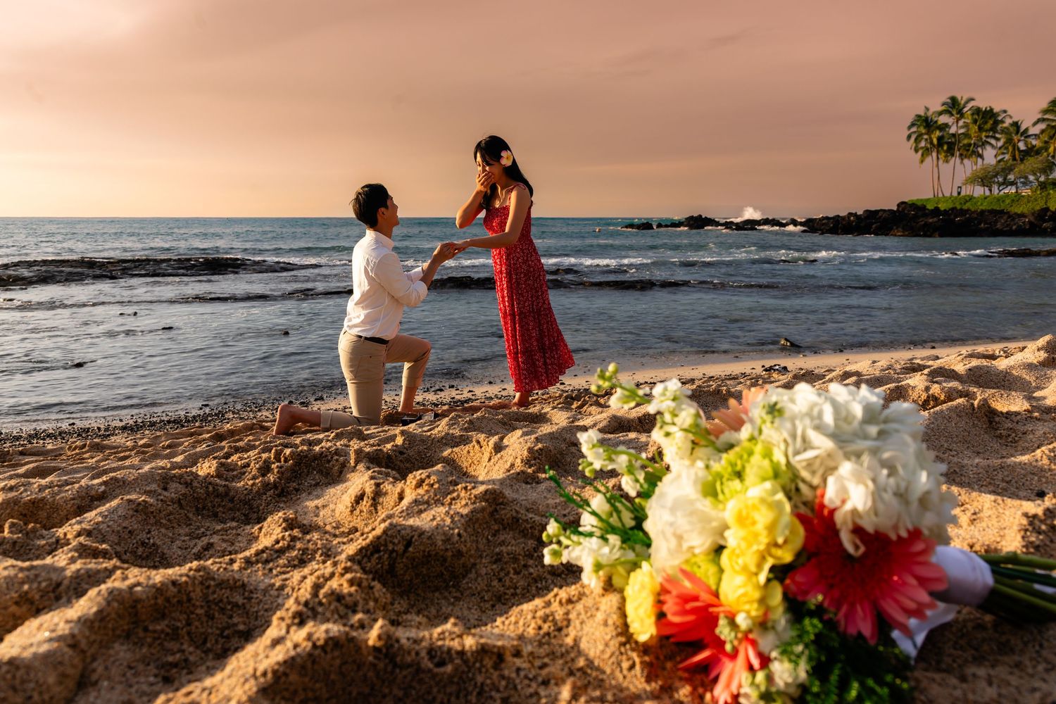 Beach proposal at sunset with colorful flowers in the foreground at Kukio beach in Kailua-Kona, Hawaii.
