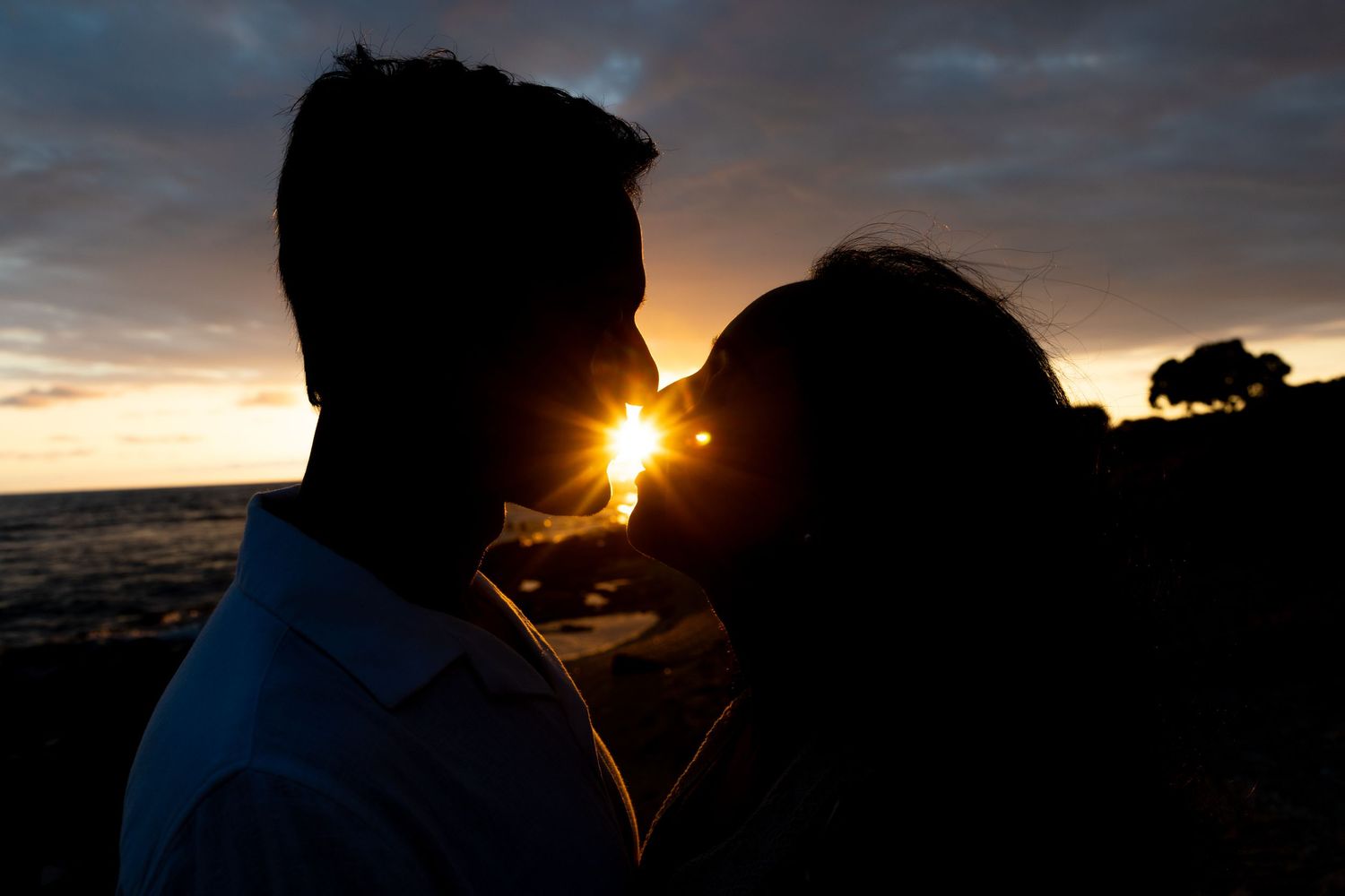 Romantic couple silhouetted against a stunning ocean sunset, sharing a kiss with golden light glowing between them.