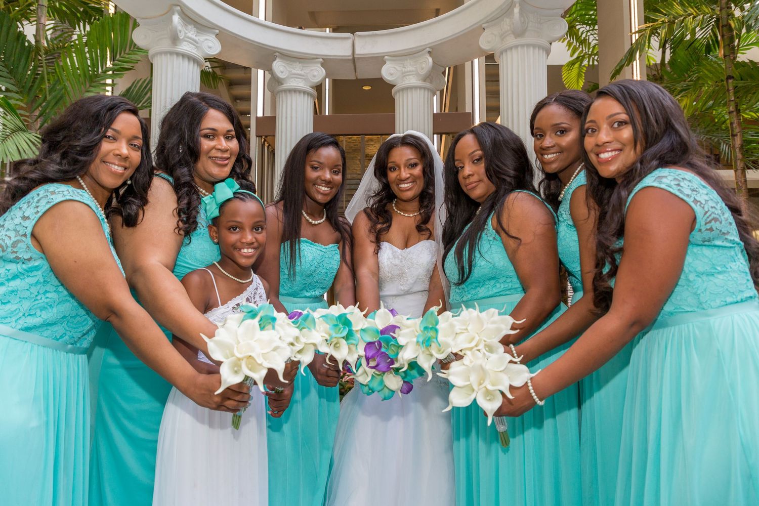 Bride in white gown poses with seven bridesmaids in teal dresses holding white bouquets at an outdoor wedding venue.