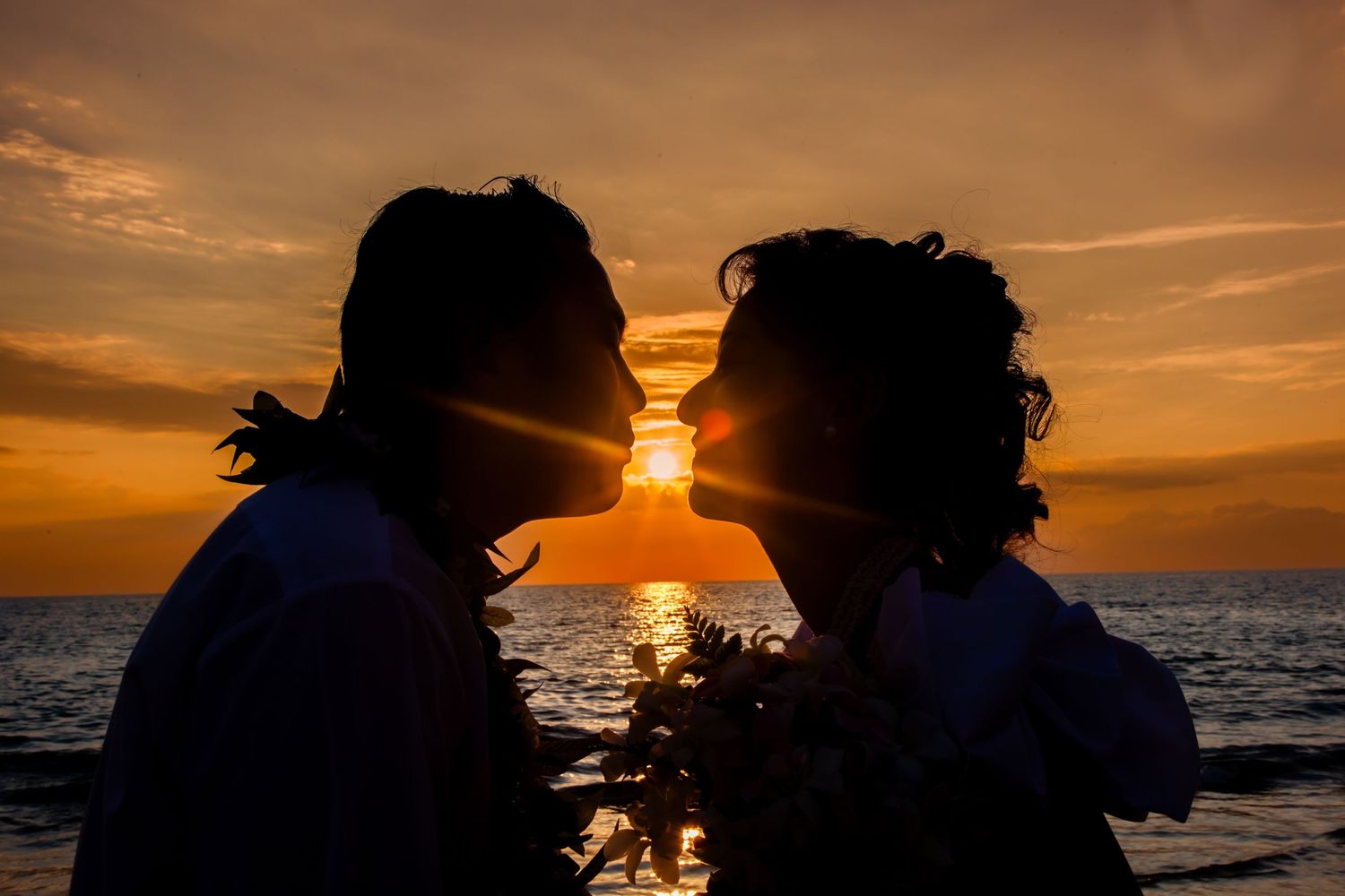 A silhouetted couple shares a romantic kiss against a vibrant orange sunset over the ocean.