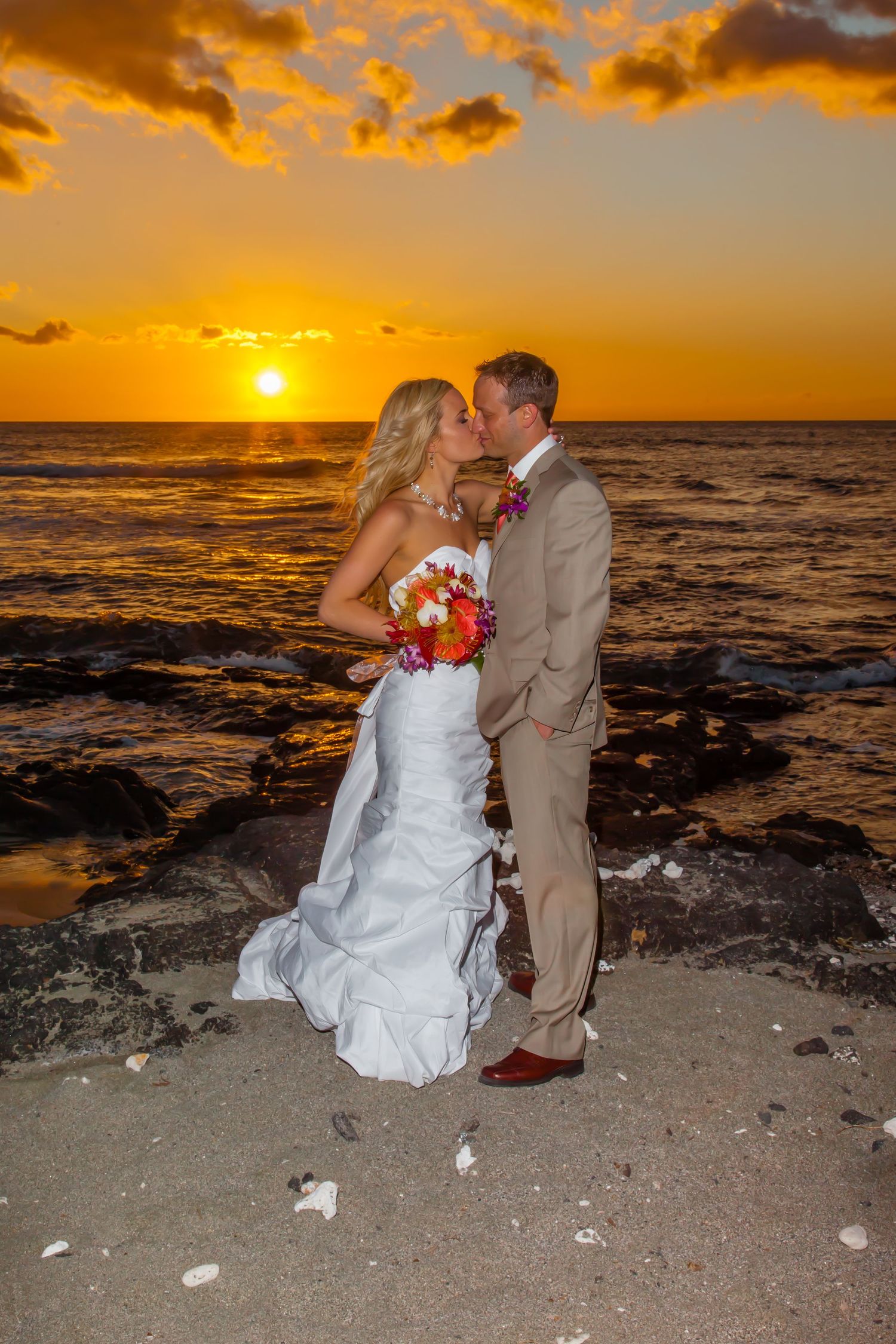 A wedding couple against a vibrant orange sunset over the ocean at the Mauna Lani beach near Kailua Kona, Hawaii.