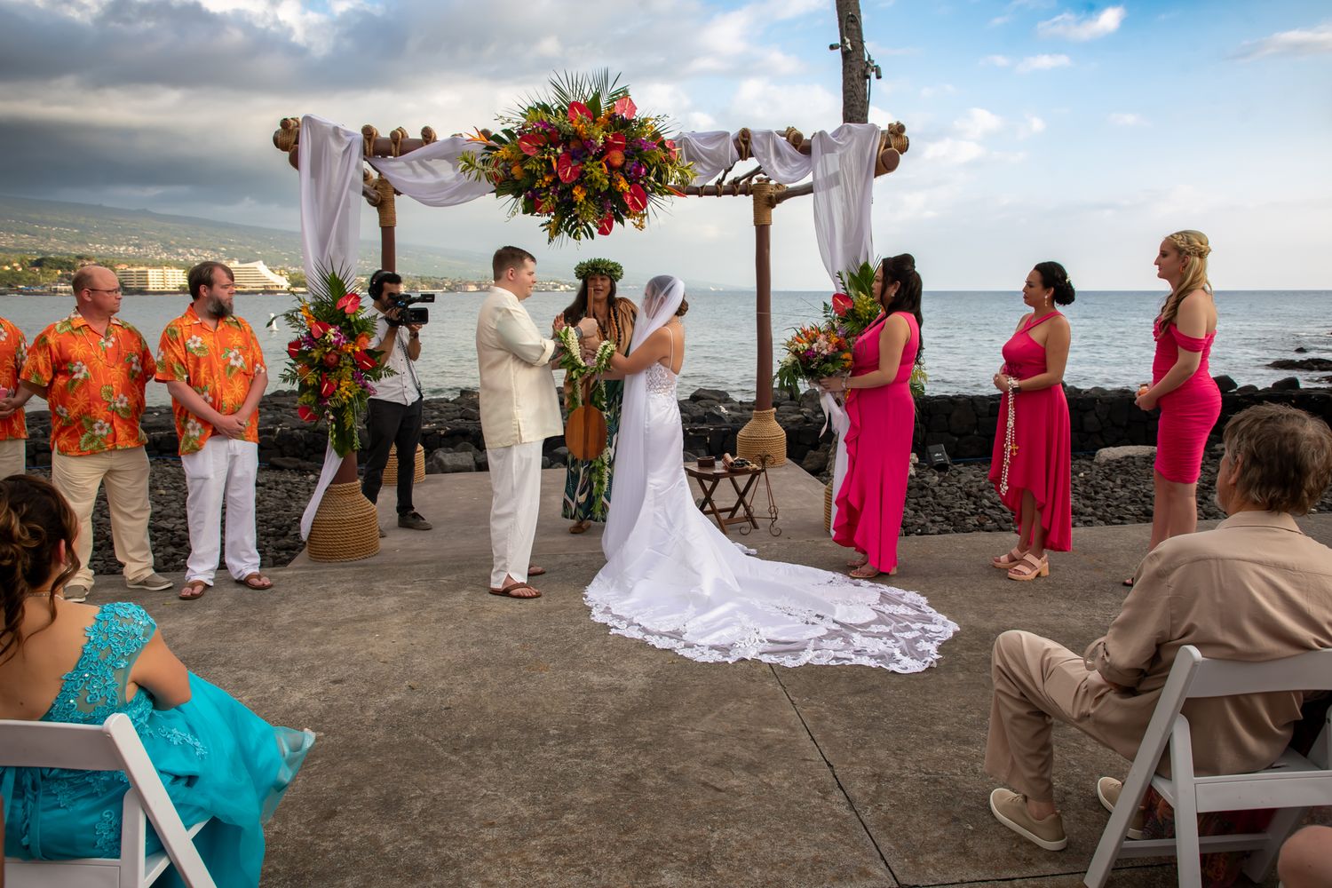 Seaside wedding ceremony under a floral arch with wedding party in orange and pink attire beside the ocean.