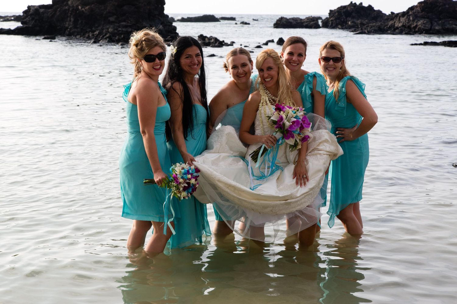 Bridesmaids in aqua dresses and bride in white gown stand together in ocean water for fun beach wedding photos.