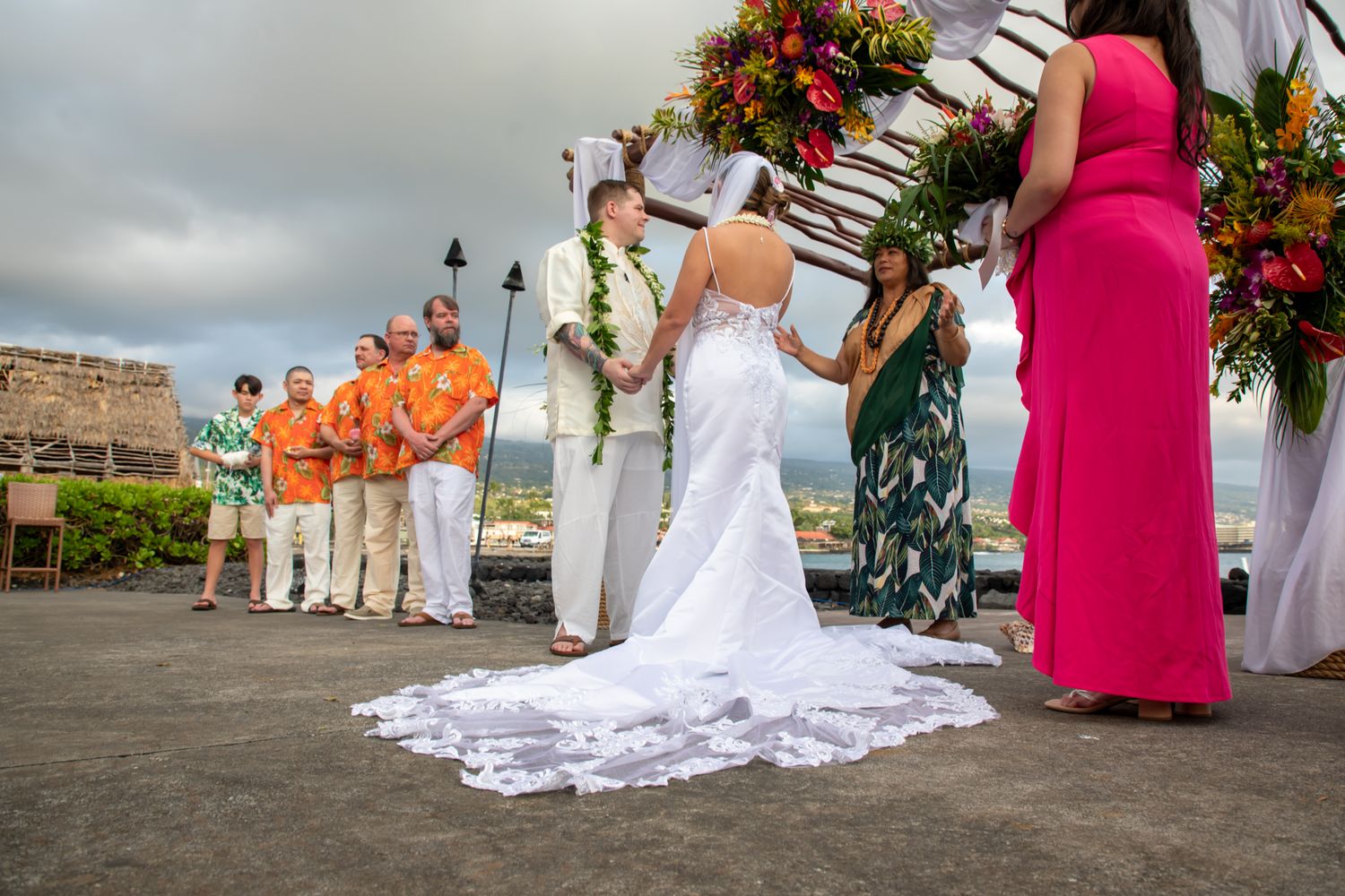 Couple exchanging vows at outdoor Hawaiian wedding ceremony with floral arch, guests in orange shirts, ocean view backdrop.