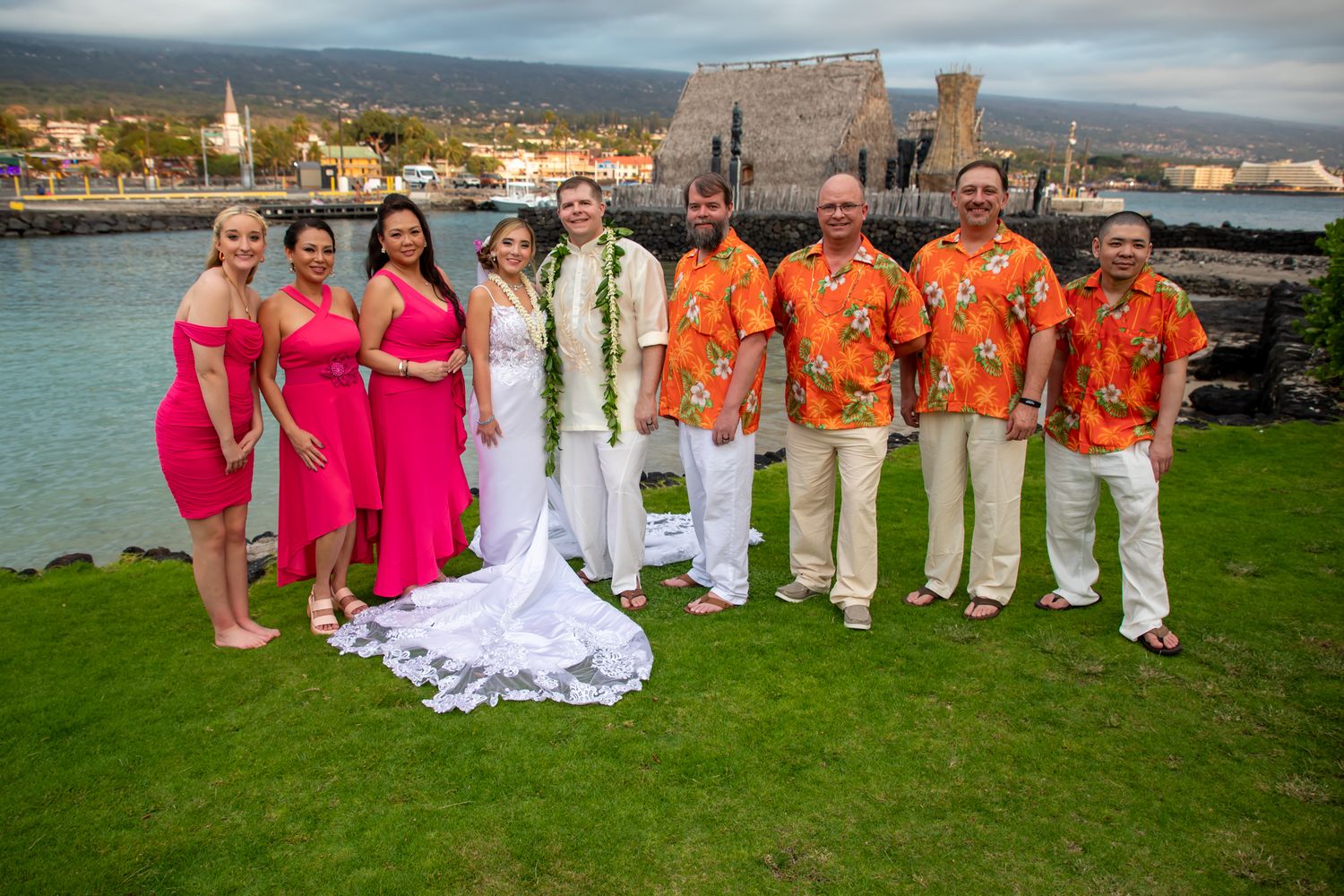 Wedding party in bright pink dresses and orange aloha shirts pose on oceanfront lawn in Hawaii.