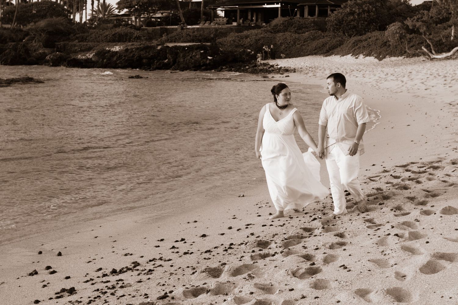 A couple in white attire walks hand in hand along a sepia-toned tropical Hawaiian beach leaving footprints in the sand.