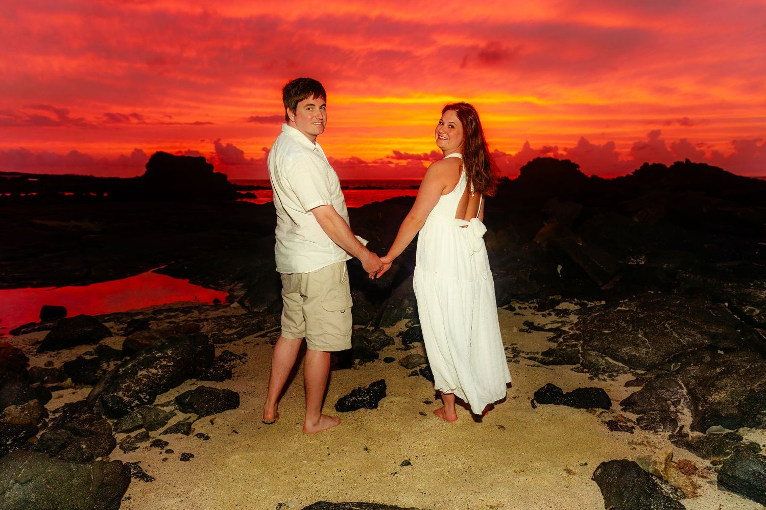 A couple holds hands on a rocky beach at sunset with dramatic red and orange sky above them.