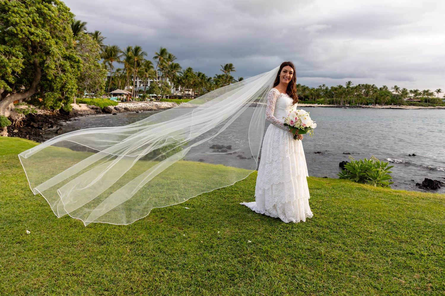Bride with flowing veil poses by oceanfront with bouquet on grassy Hawaiian shoreline.