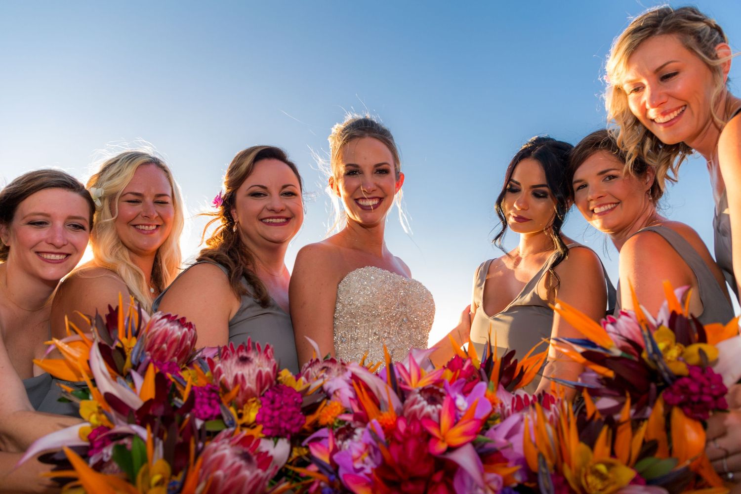 Bride and bridesmaids in gray dresses smiling together holding vibrant pink and orange tropical flower bouquets outdoors.