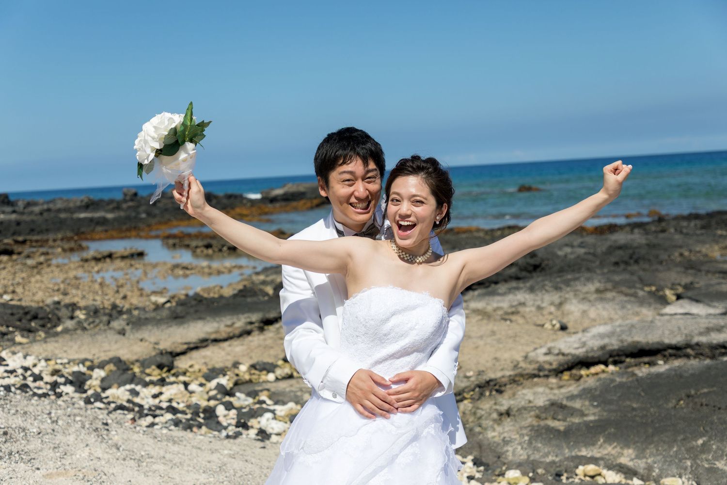 Happy newlywed couple celebrating on a rocky beach, bride in white strapless gown holding bouquet, groom in white shirt.