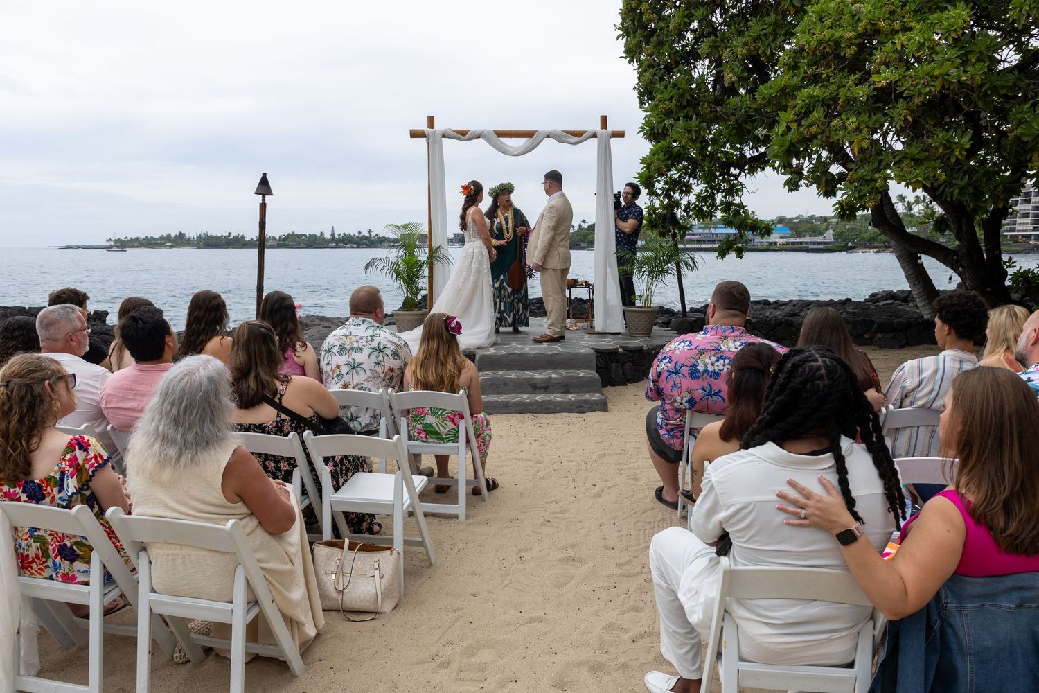 Outdoor beach wedding ceremony with guests seated on white chairs facing couple at floral arch by ocean.