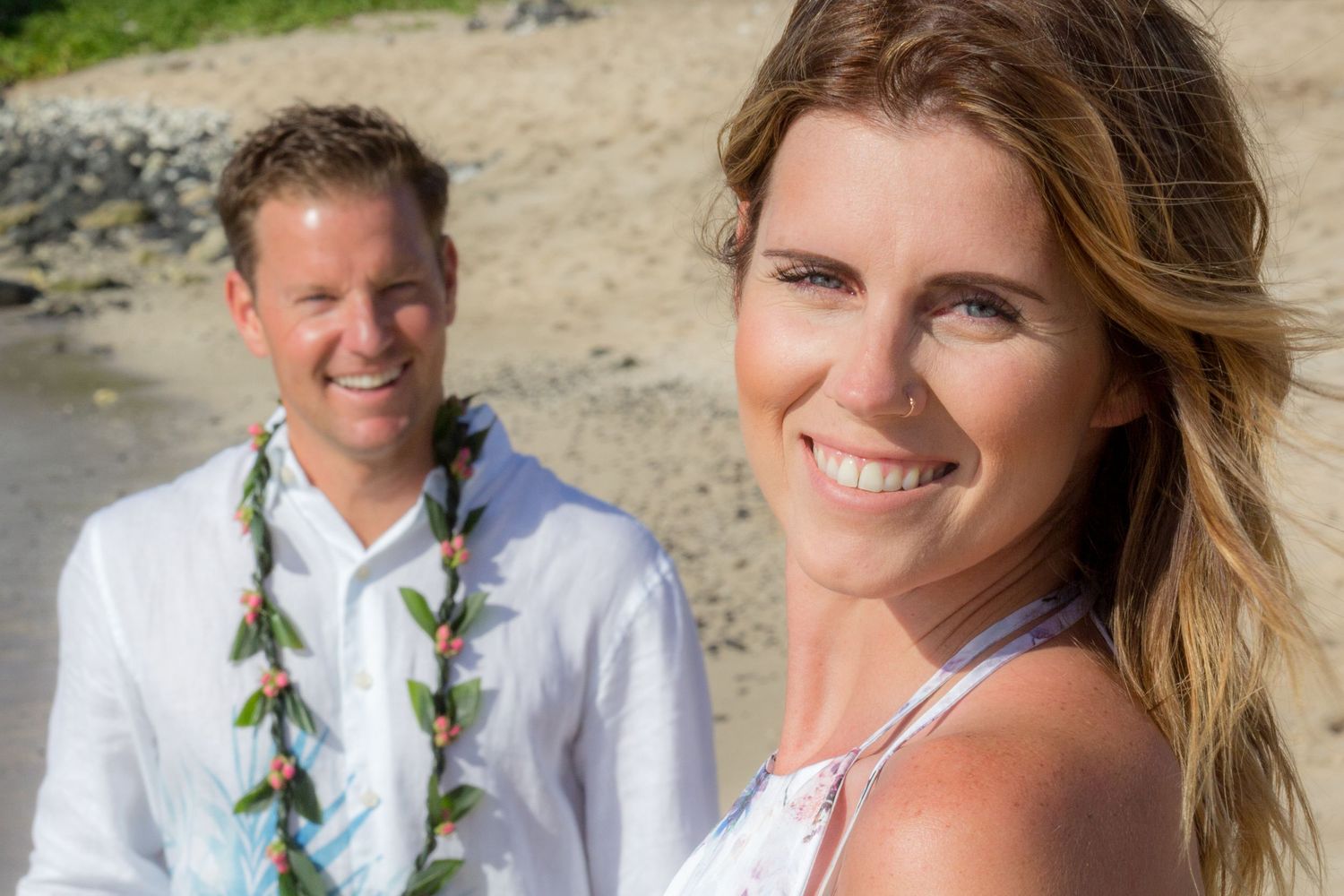 Couple in Hawaiian leis smiling on a tropical beach, woman in foreground wearing white, man in white shirt behind her.