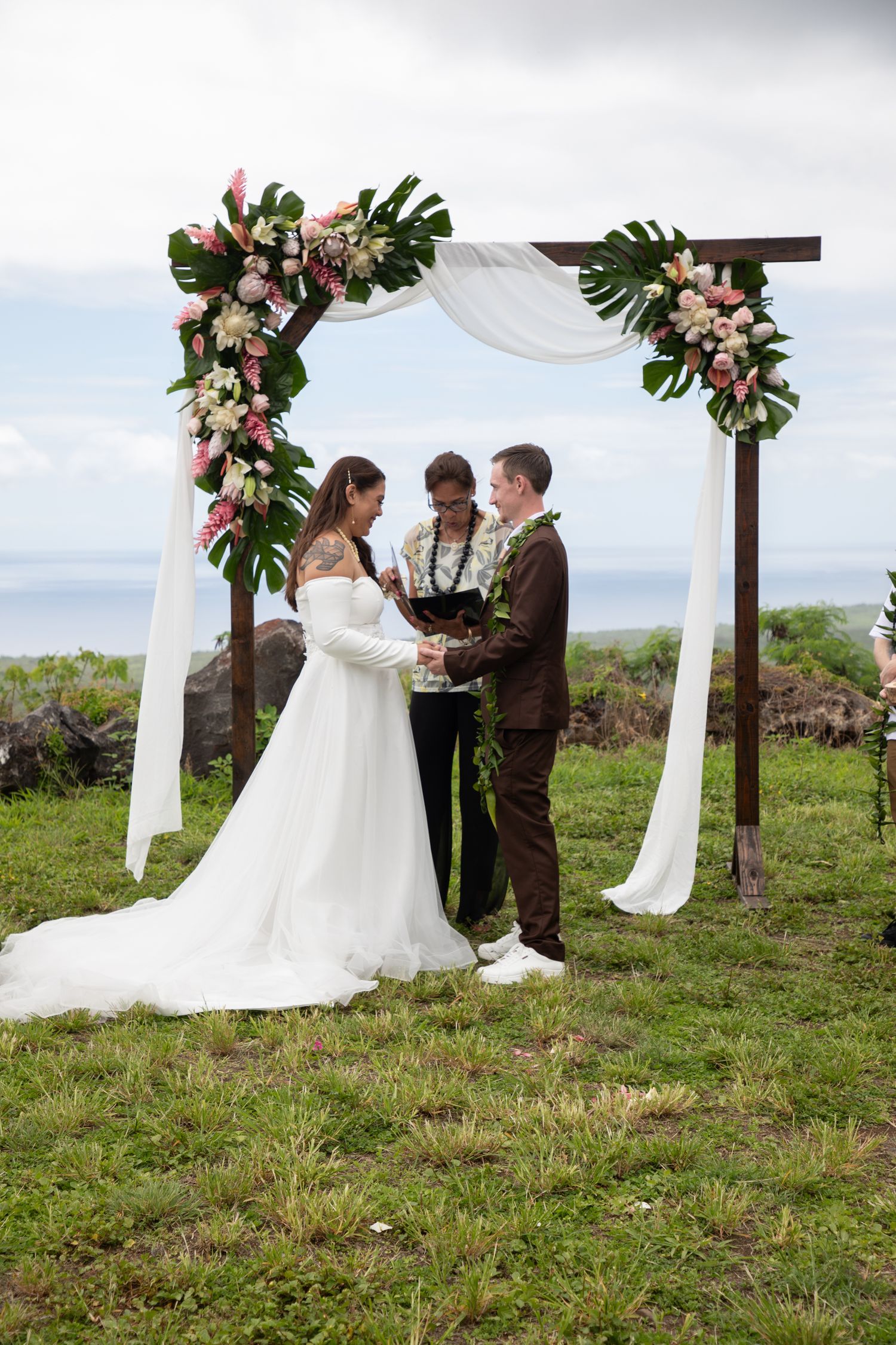 Couple exchanges vows under floral arch with white draping at outdoor oceanview wedding ceremony in Hawaii.